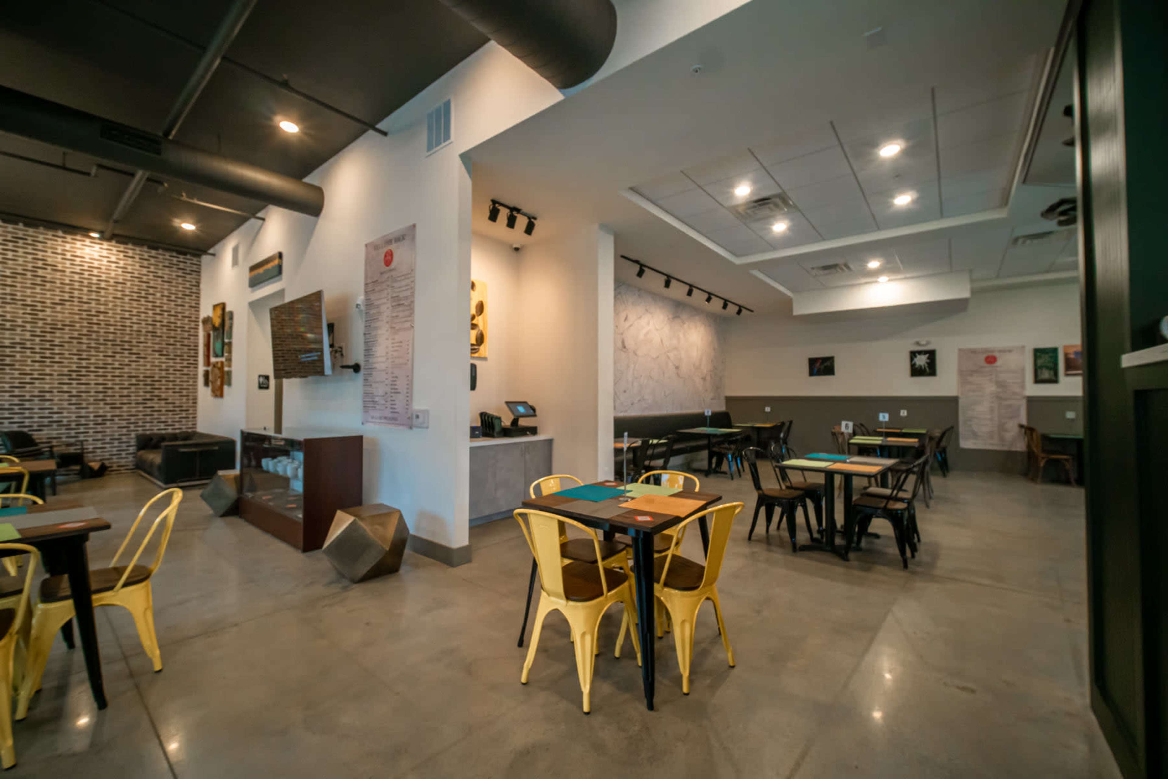 A modern restaurant interior with concrete floors, yellow chairs, wooden tables, and a textured wall featuring art.