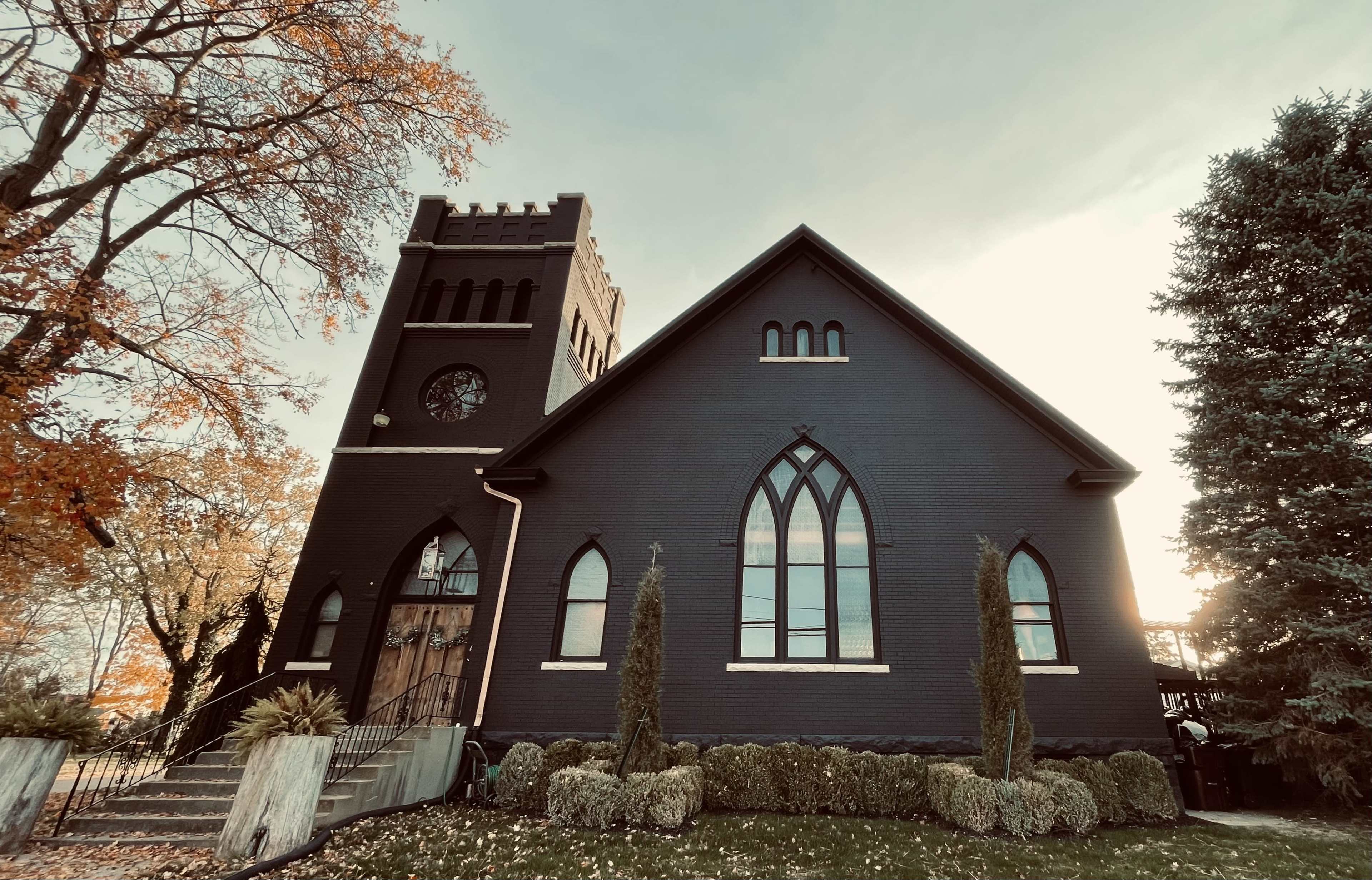 A dark-bricked church with a tall clock tower features large arched windows and is surrounded by neatly trimmed hedges and autumn foliage.