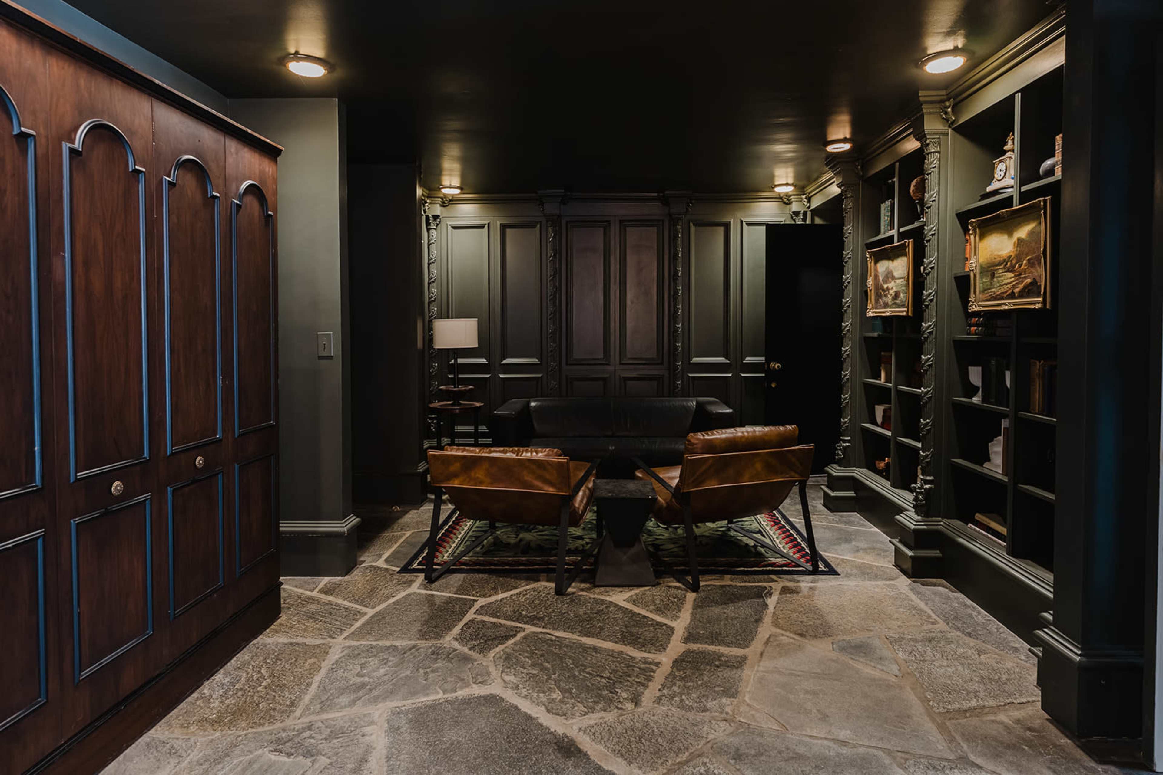 The image shows a dimly lit room featuring a pair of leather chairs and a black sofa, flanked by built-in bookshelves and a large wooden wardrobe.