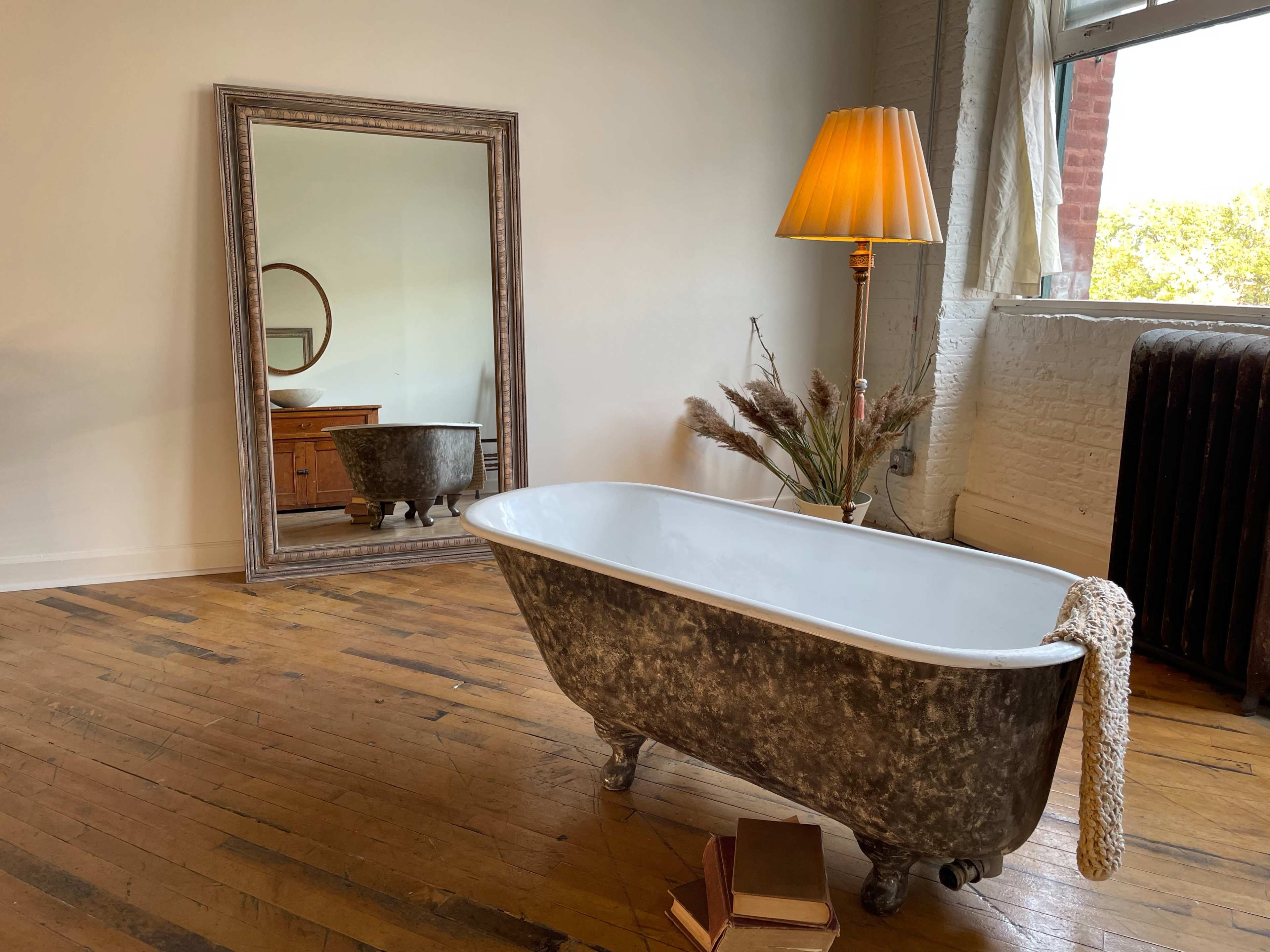 A vintage-style freestanding bathtub sits on wooden flooring beside a lamp and a large mirror in a bright, minimalist room.
