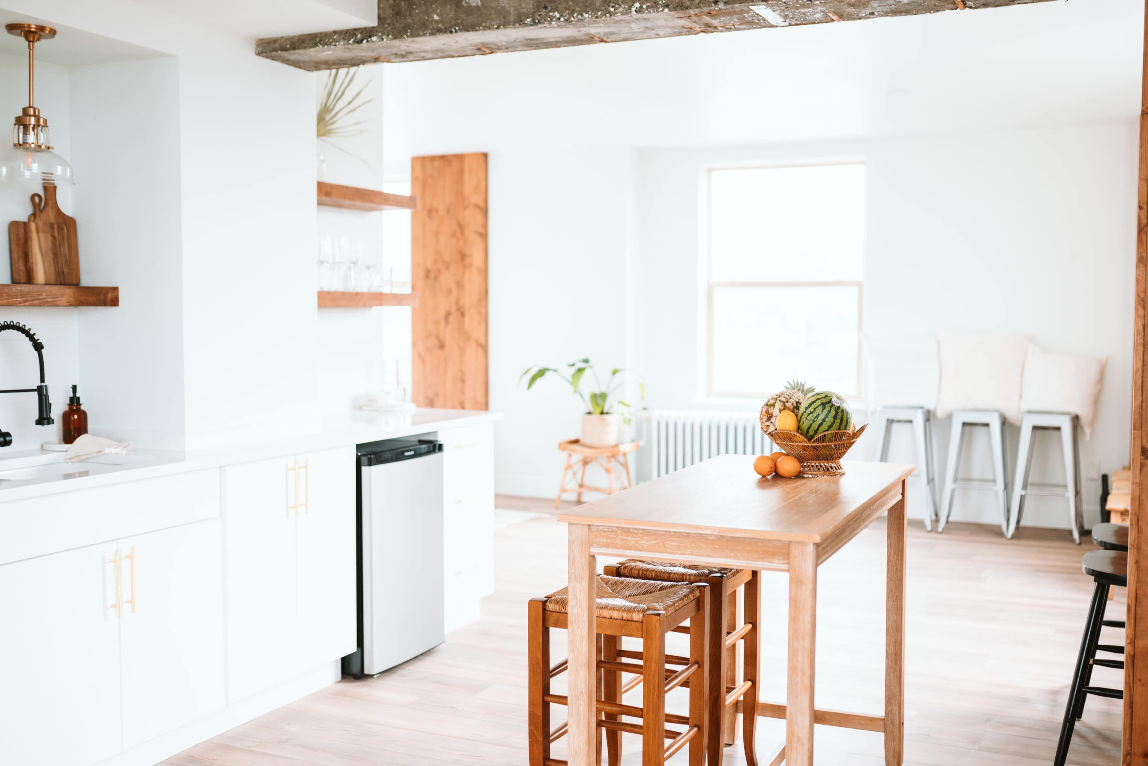 A bright kitchen features a wooden table surrounded by stools, with a fruit bowl atop the table and modern cabinetry in the background.
