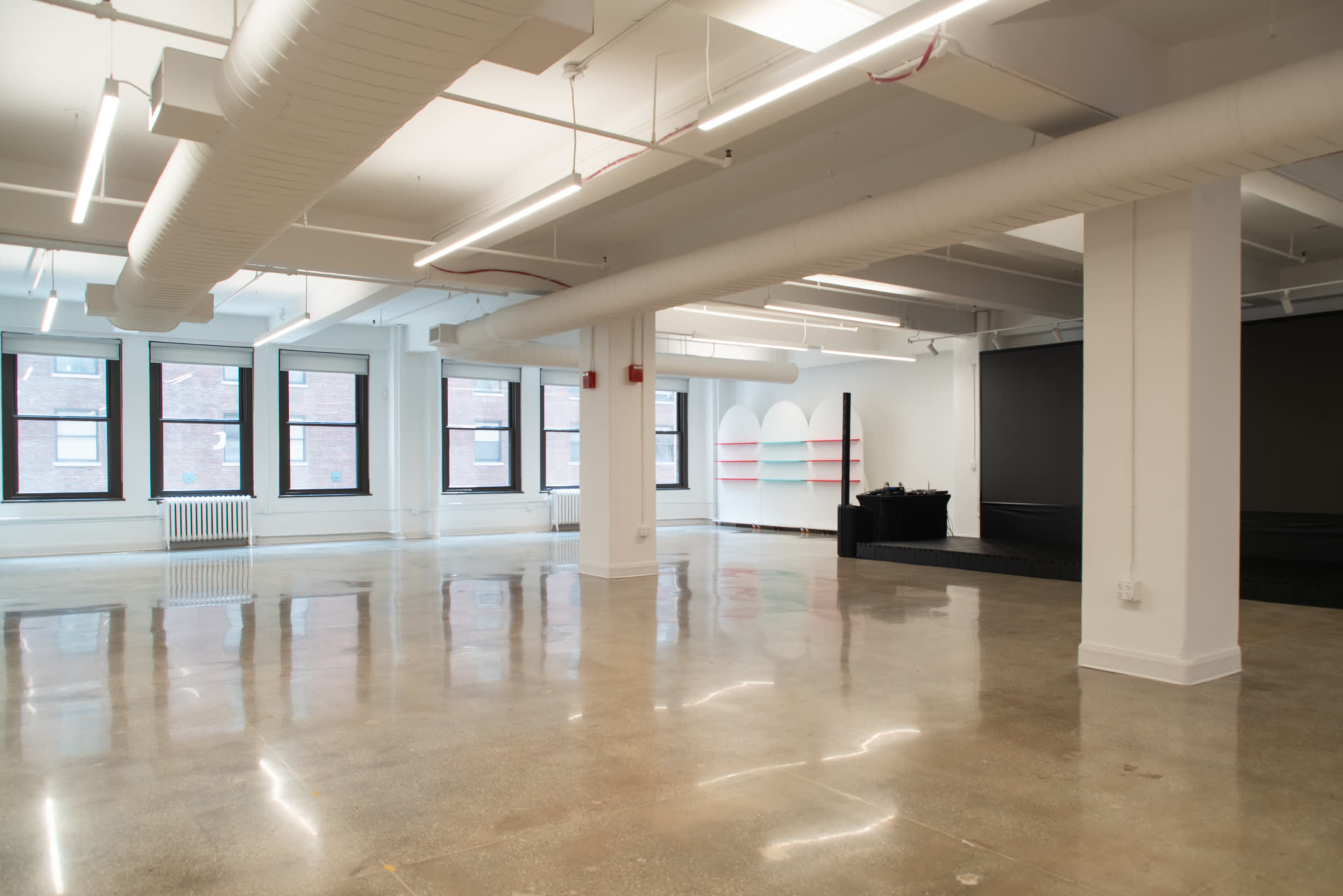 The image shows a spacious, empty room with polished concrete flooring, large windows, and simple white walls, featuring a small stage area.
