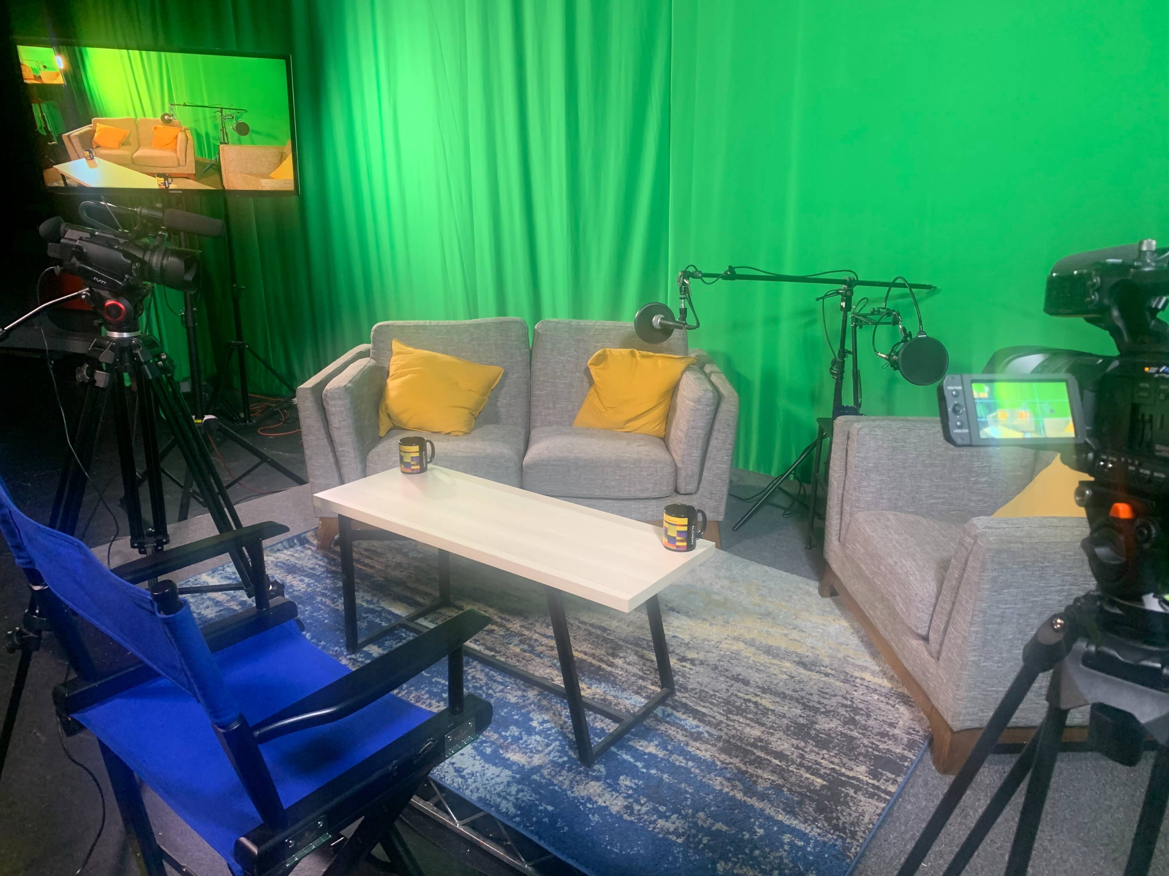A television studio setup with a green backdrop, two couches with yellow cushions, a coffee table, and multiple cameras positioned around the area.