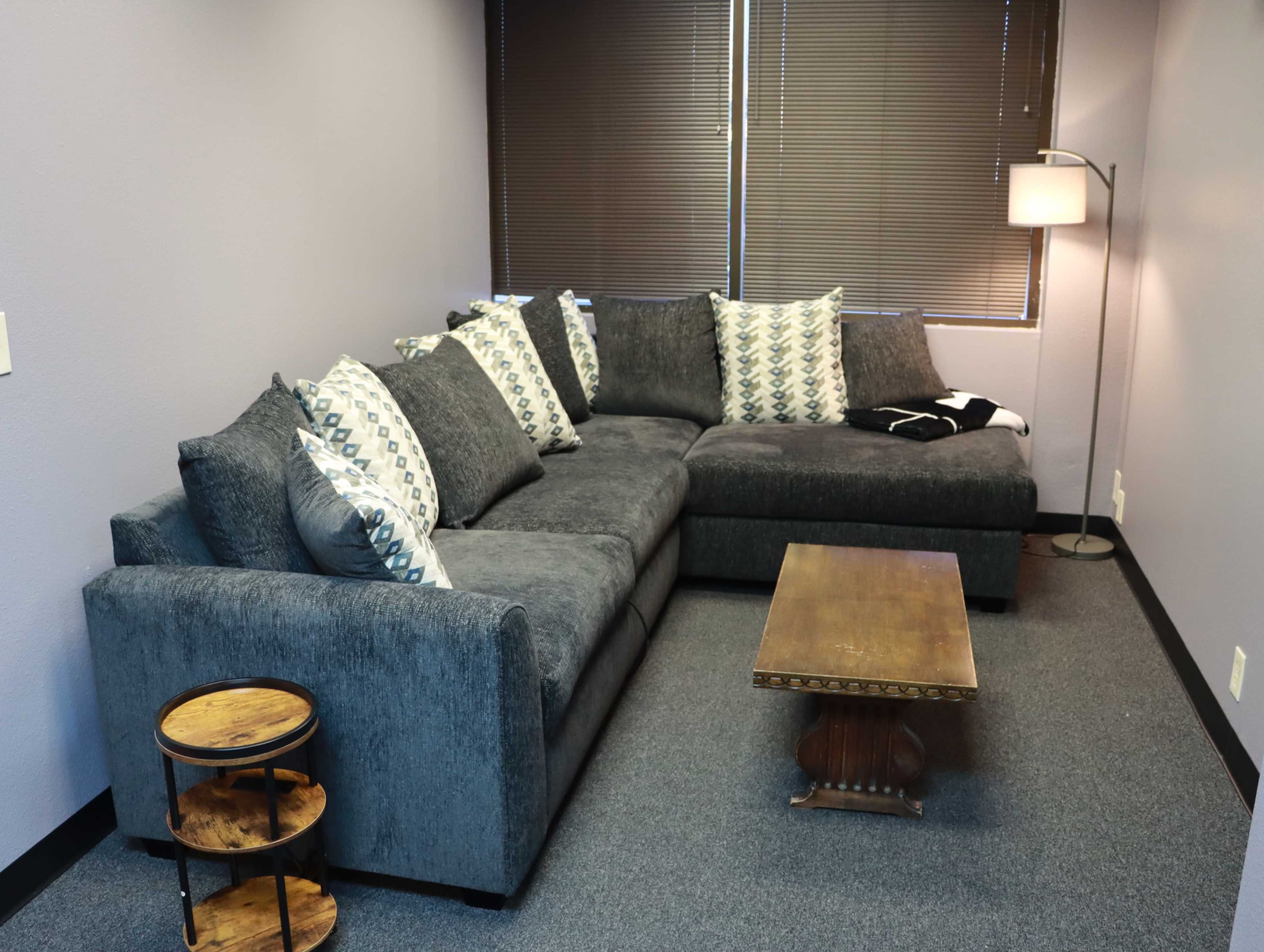 The image shows a gray sectional couch with patterned pillows, a wooden side table, and a floor lamp in a small, well-lit room with dark window blinds.