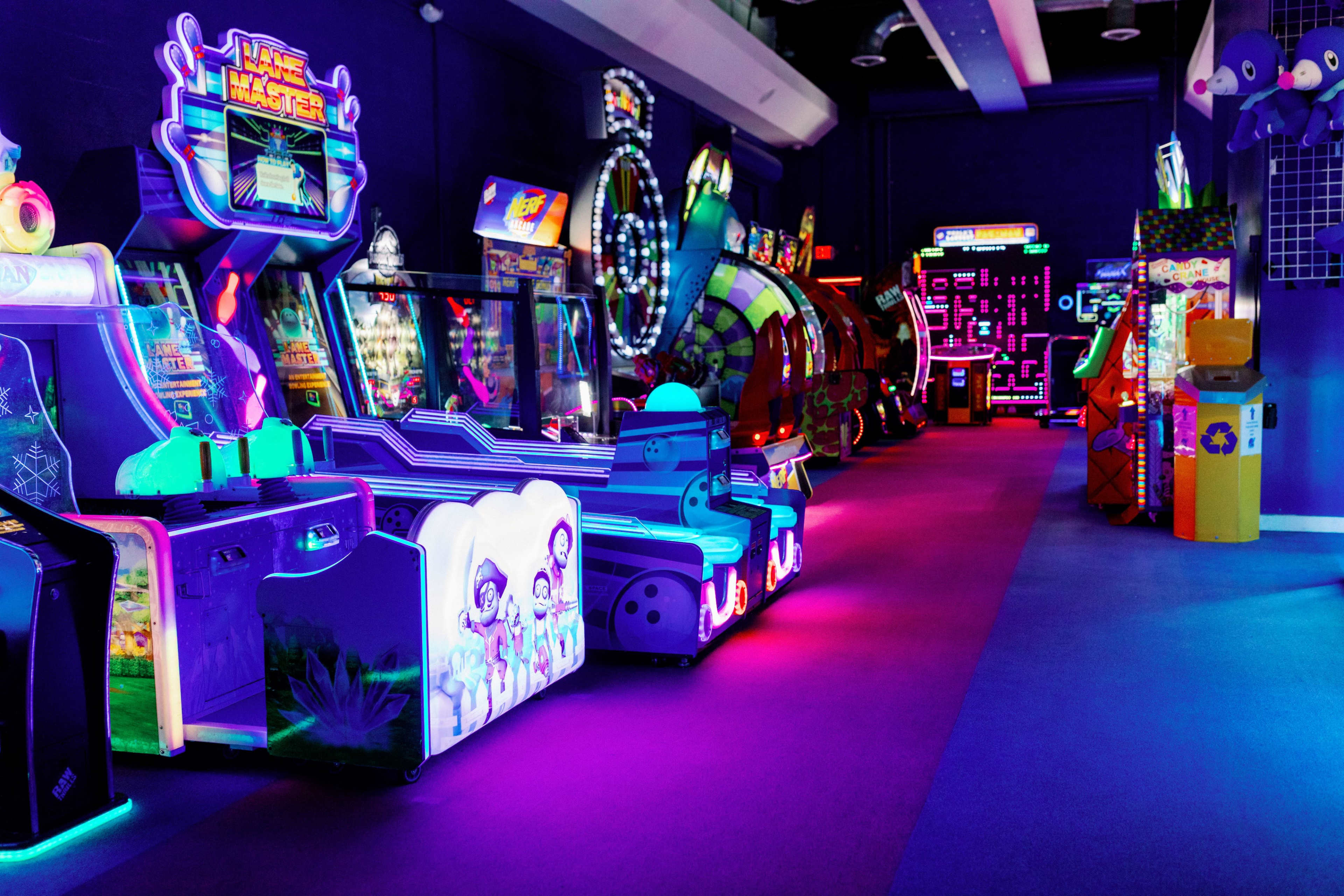 The image shows a brightly lit arcade filled with various video game machines and colorful decorations along the walls.