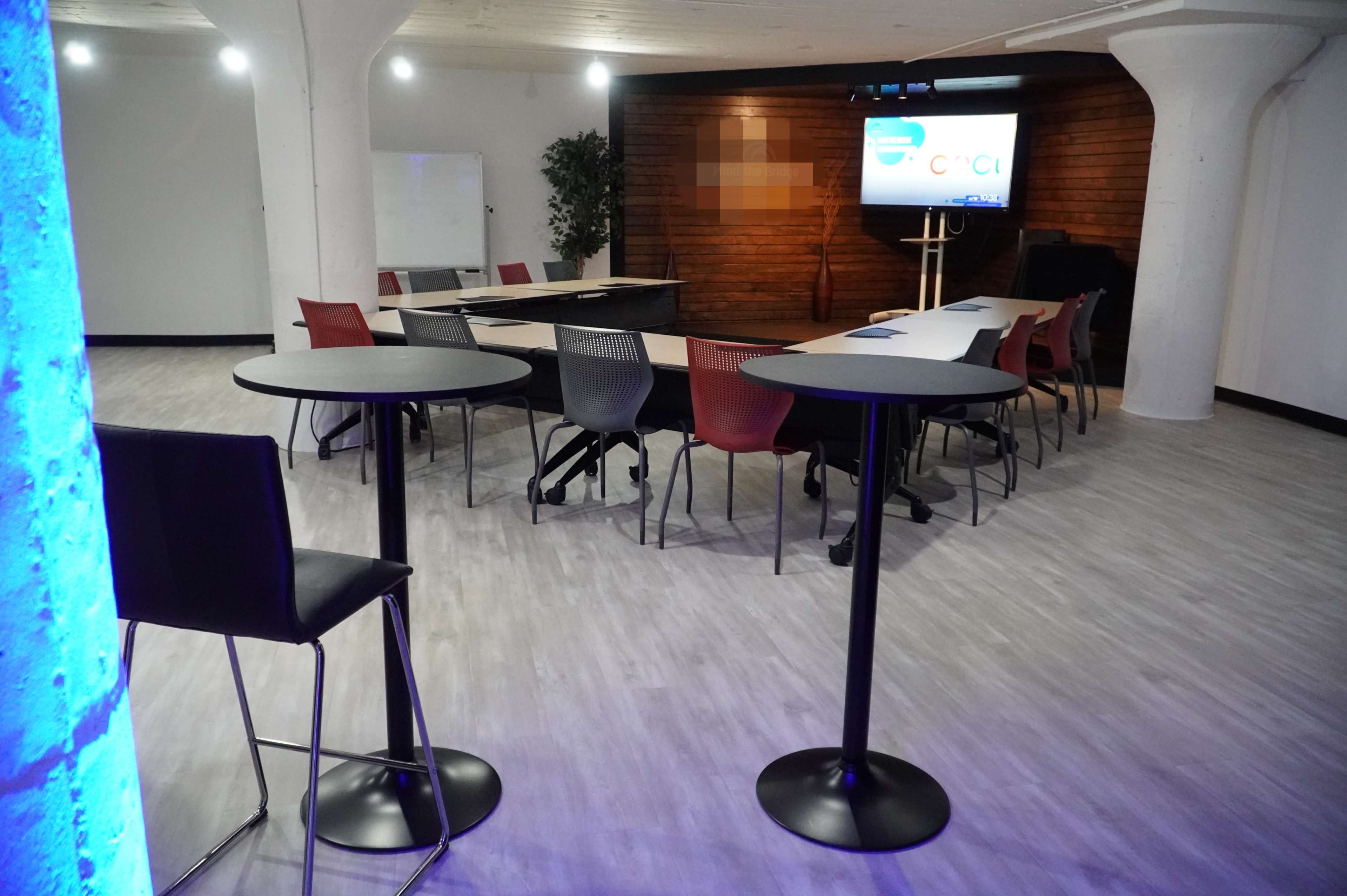 A modern meeting room with multiple tables arranged in a U-shape, surrounded by chairs, and a screen displaying a logo on the front wall.