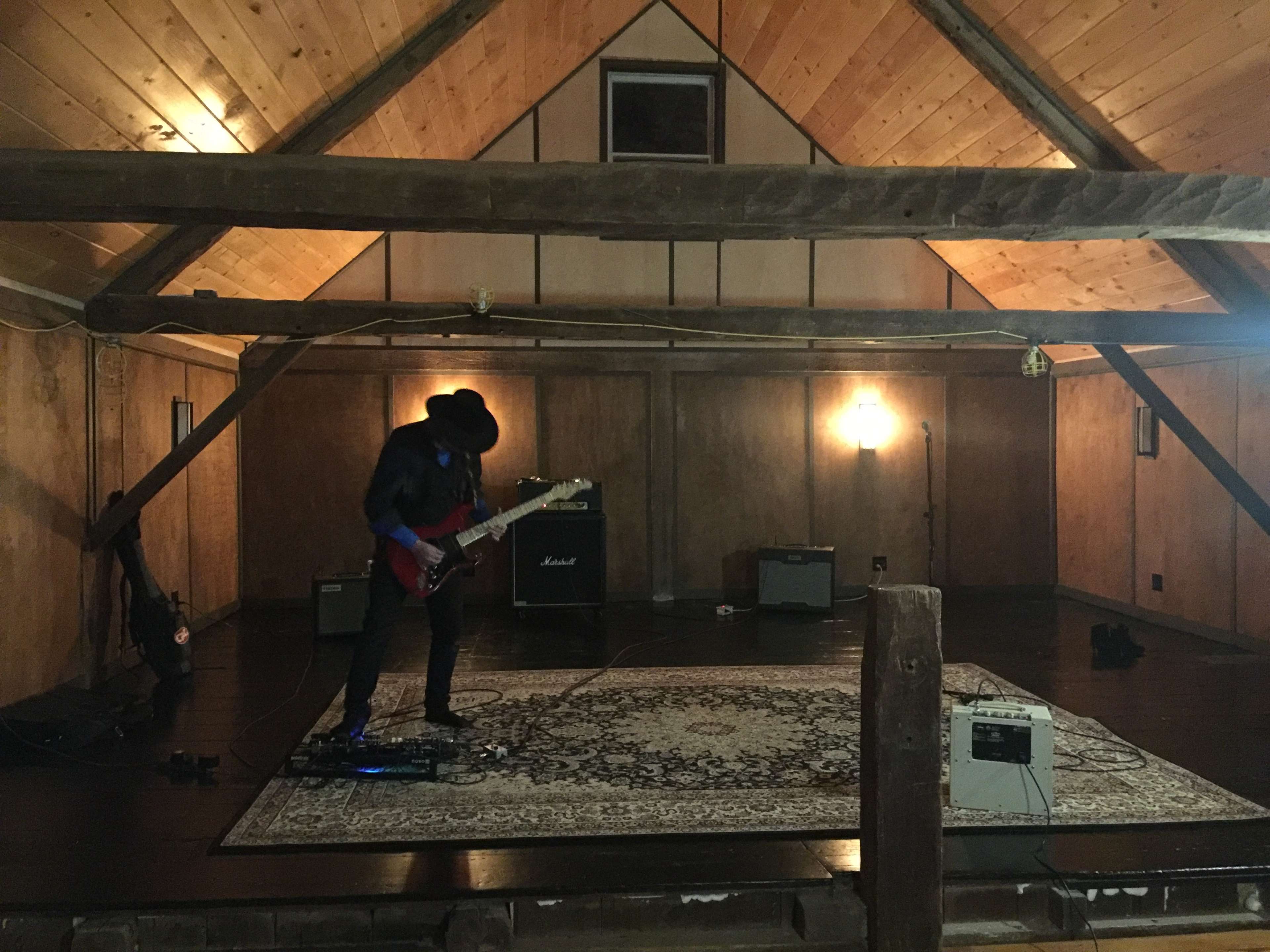 A musician stands on a rug in an empty wooden room, playing an electric guitar near amplifiers set against the walls.