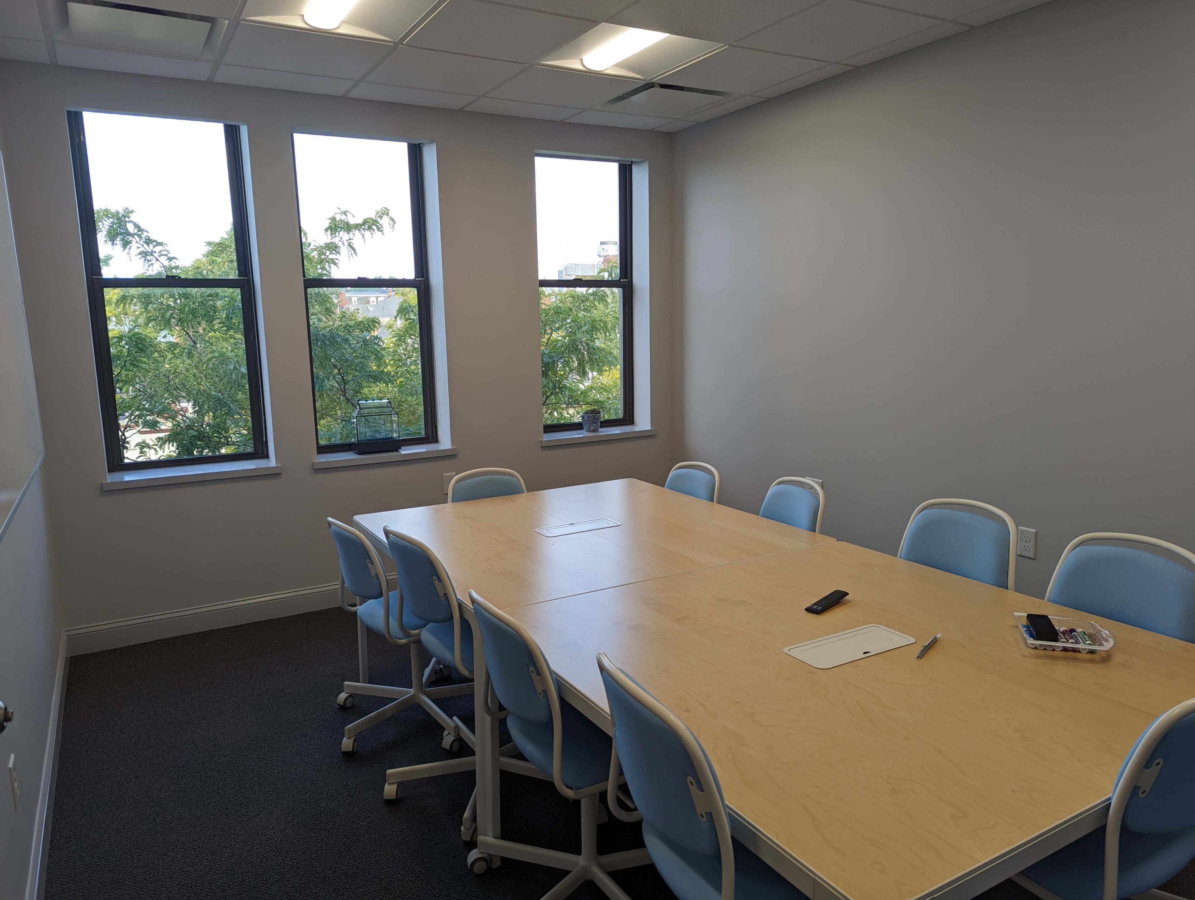 A conference room features a long wooden table surrounded by blue chairs and large windows with views of trees outside.