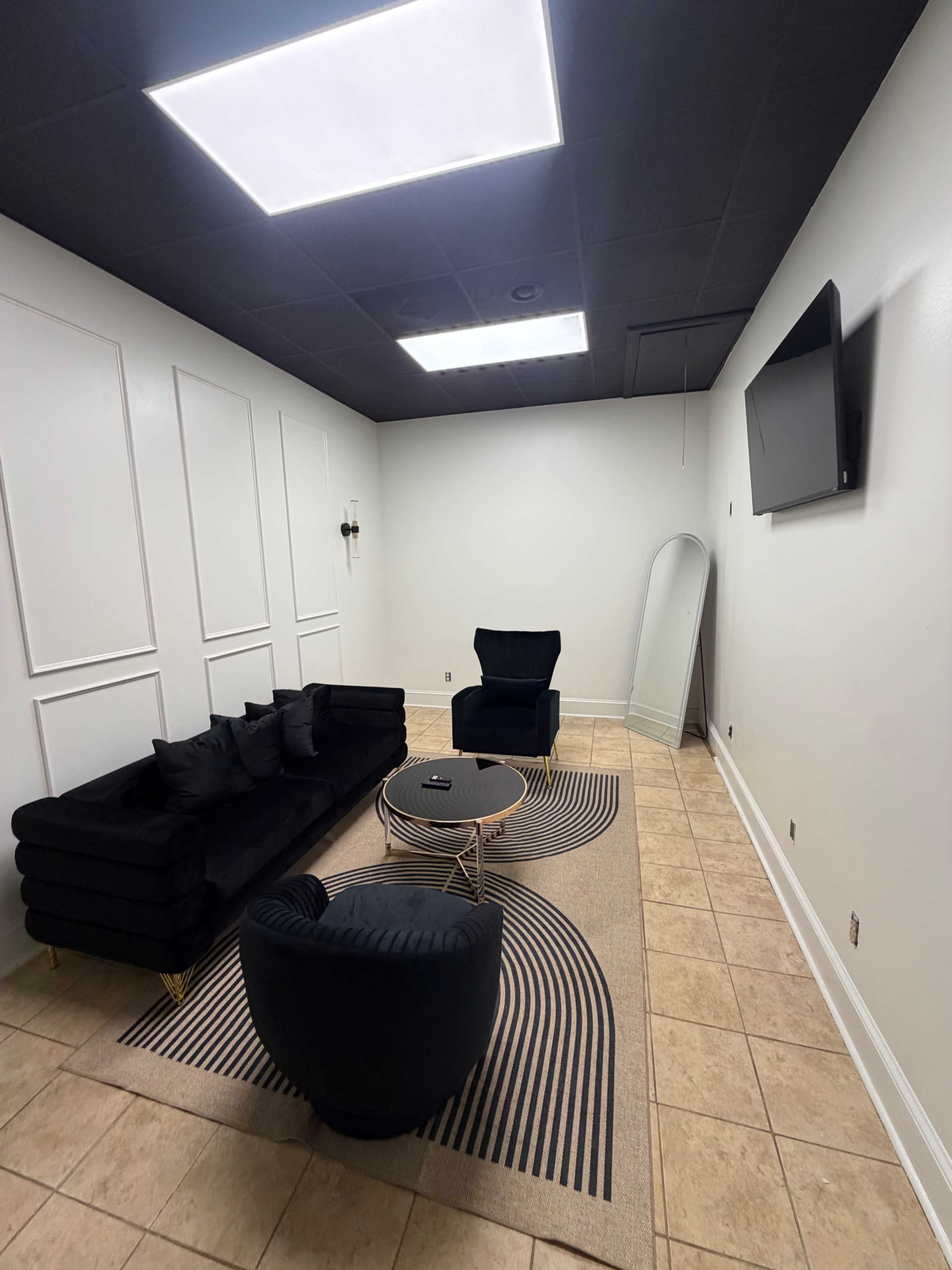 The image shows a minimally furnished room with a black sofa, a blue armchair, a round coffee table on a striped rug, a full-length mirror, and a wall-mounted TV.