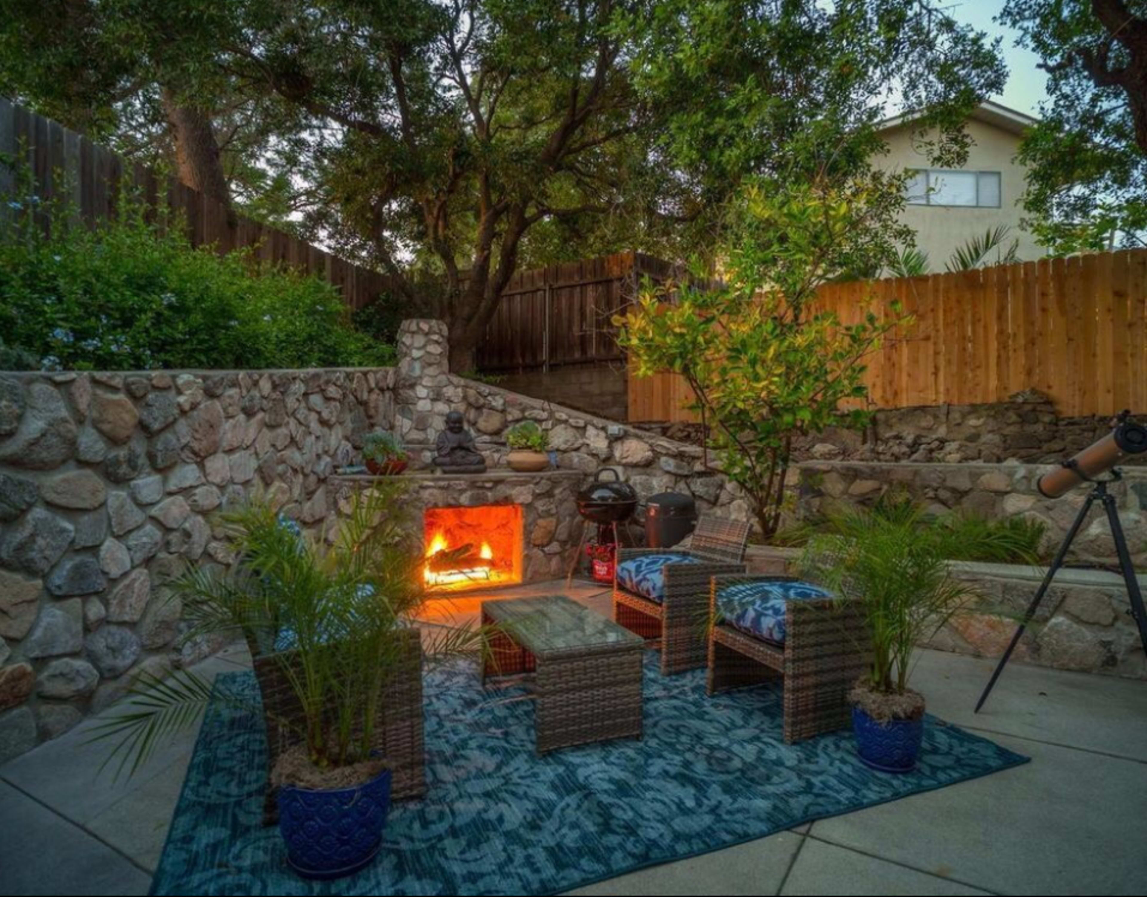 The image shows a stone patio area with outdoor furniture, a fire feature, and a telescope, surrounded by trees and decorative plants.