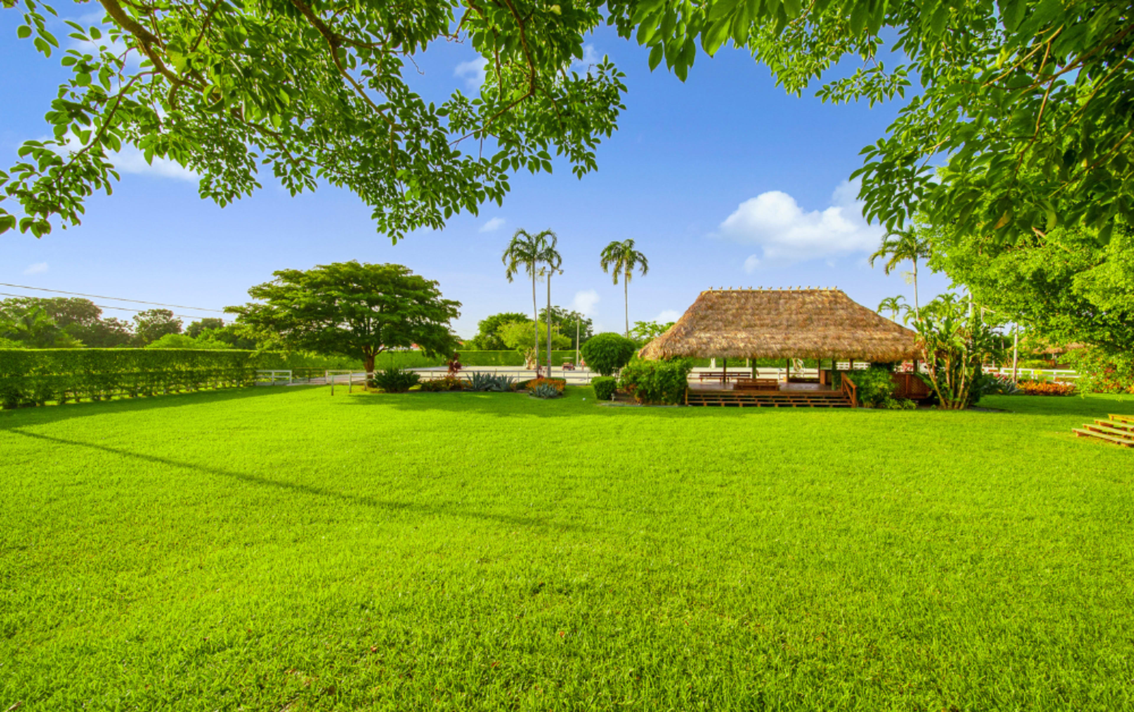 A thatched-roof gazebo sits on a lush green lawn surrounded by trees and palm plants under a clear blue sky.