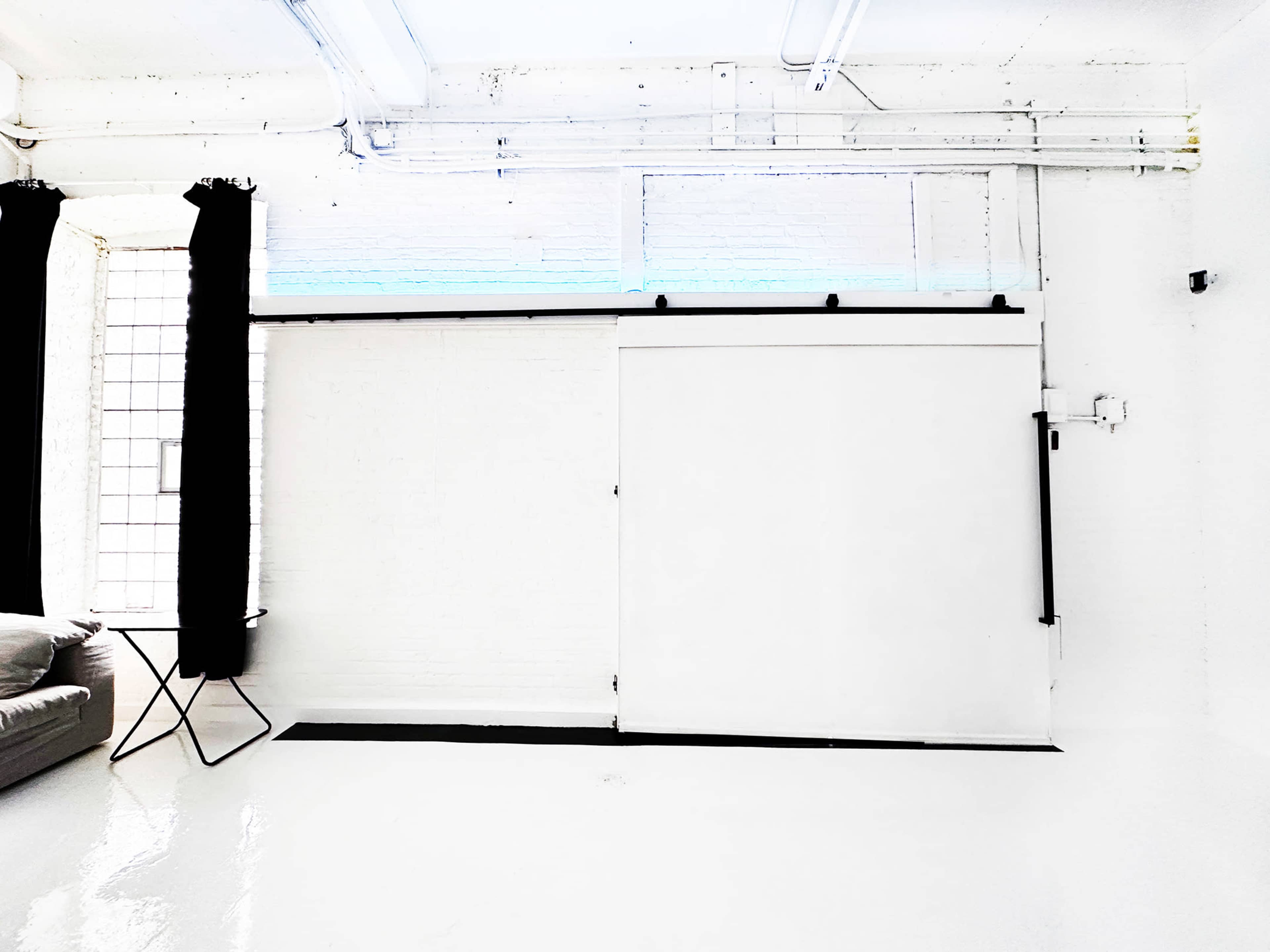 A bright, minimalistic interior with a sliding white door, a sofa, and a small table beside a window with black curtains.