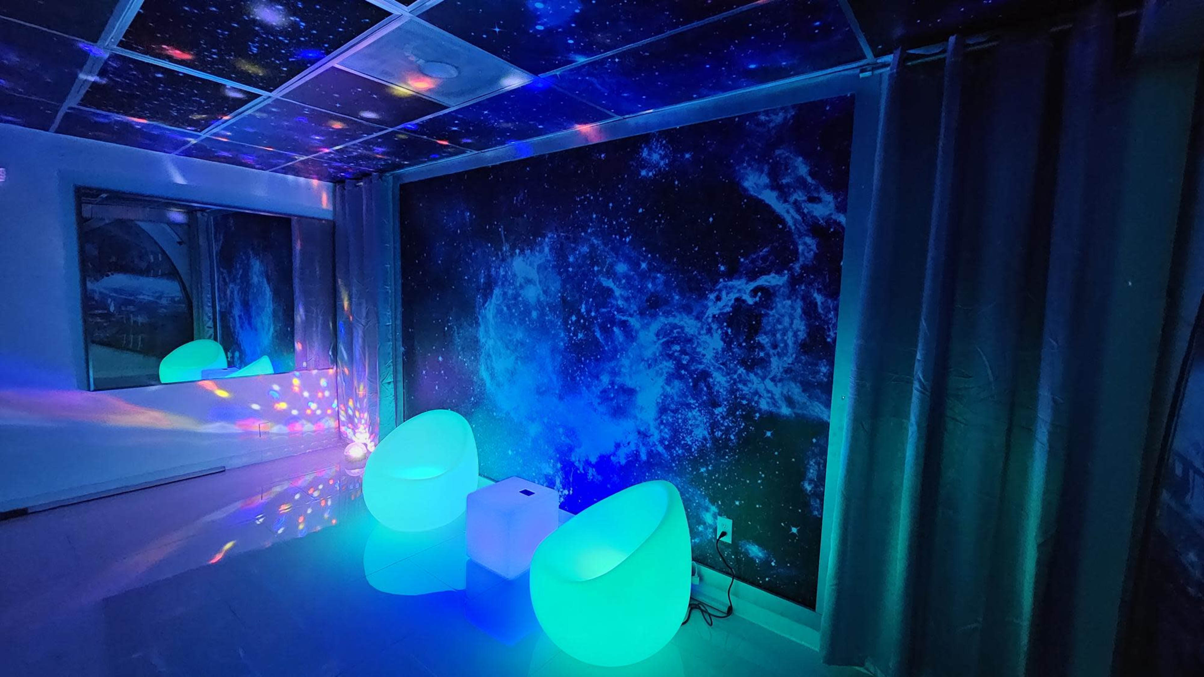 The room features a dark wall with a galaxy mural, illuminated seating, and a projector casting colorful lights across the ceiling and floor.
