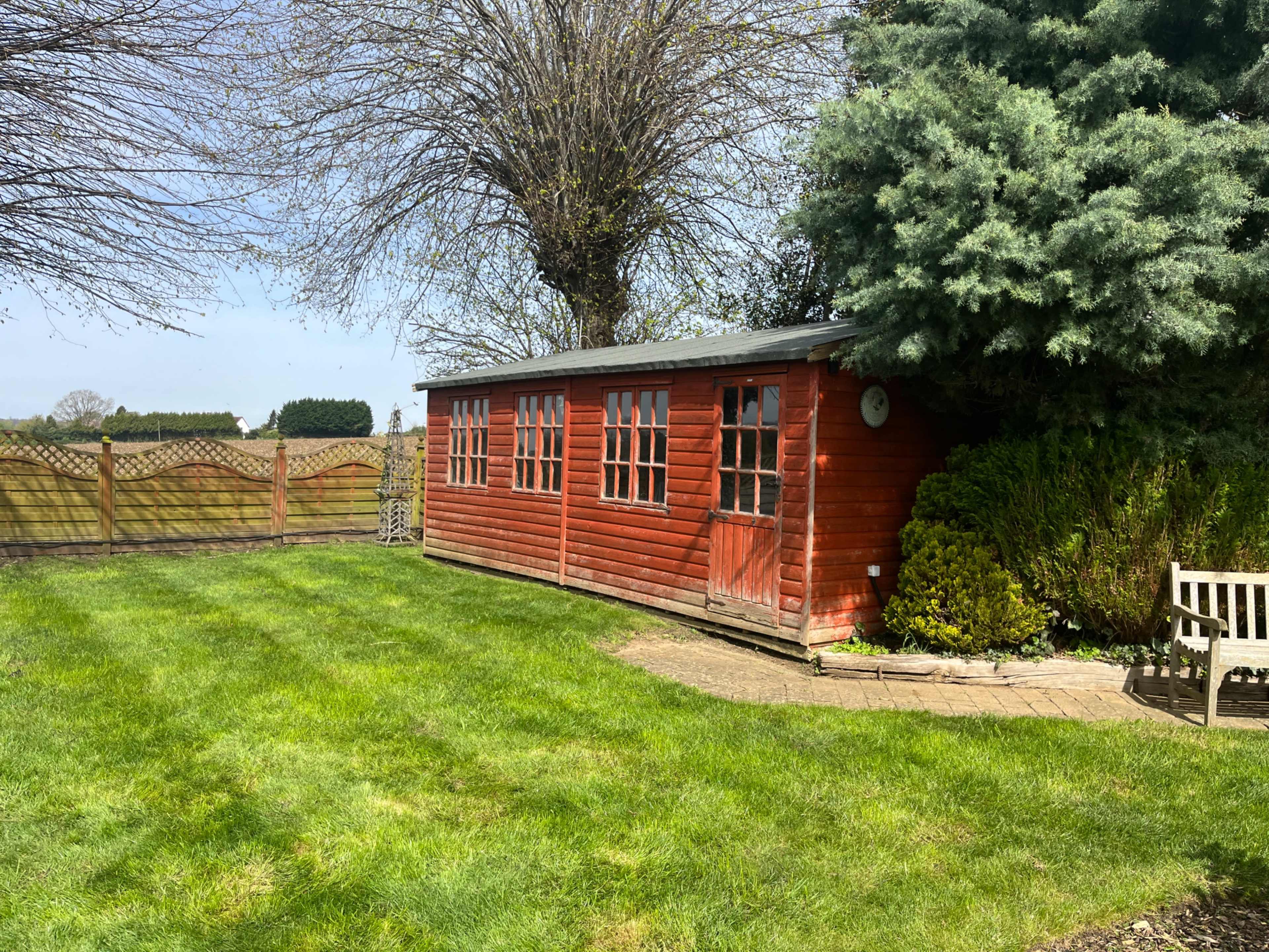 A red wooden shed with multiple windows stands in a grassy yard surrounded by trees and a wooden fence.
