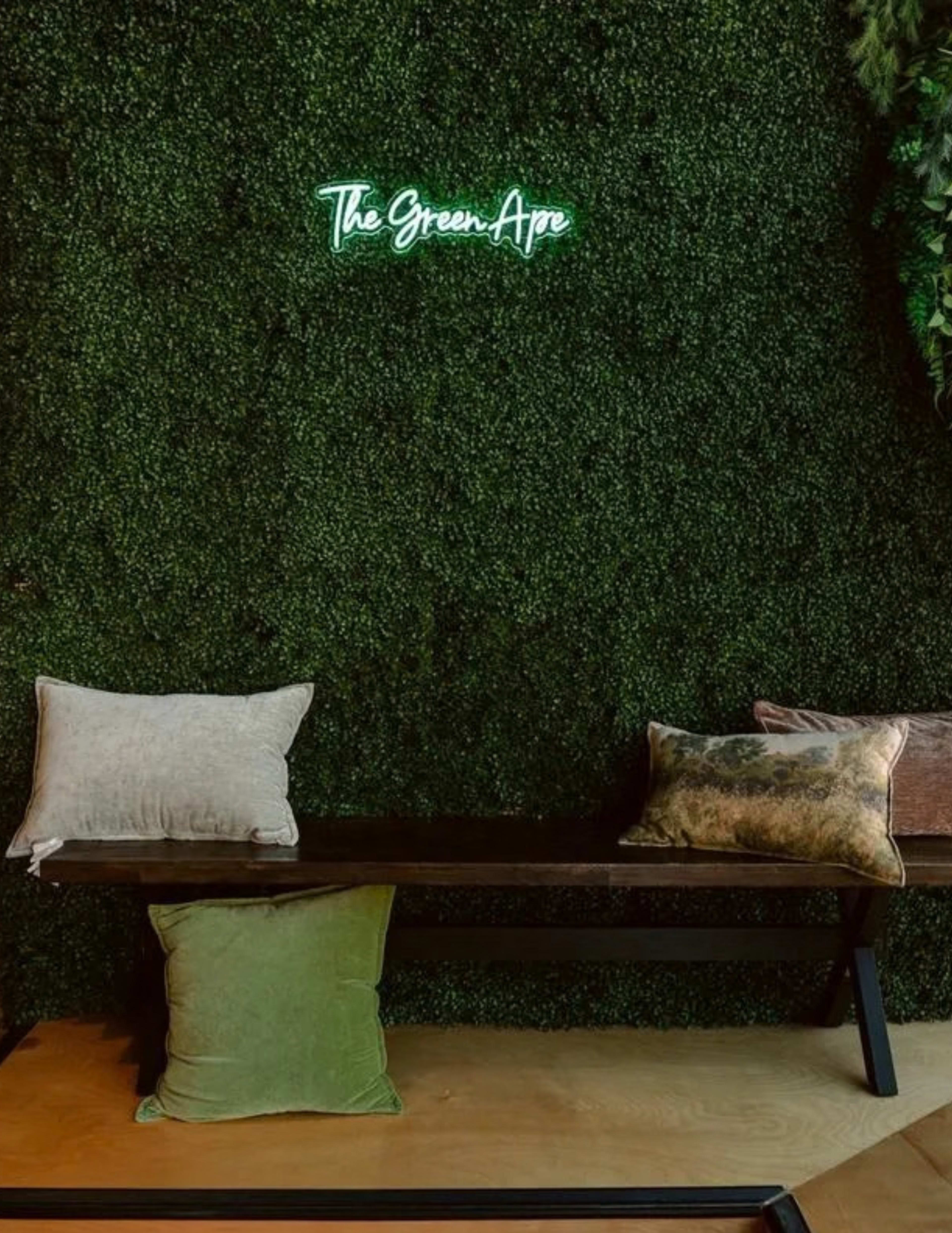 The image shows a wooden bench with cushions in front of a green moss-covered wall, showcasing a neon sign that reads "The Green Ape."