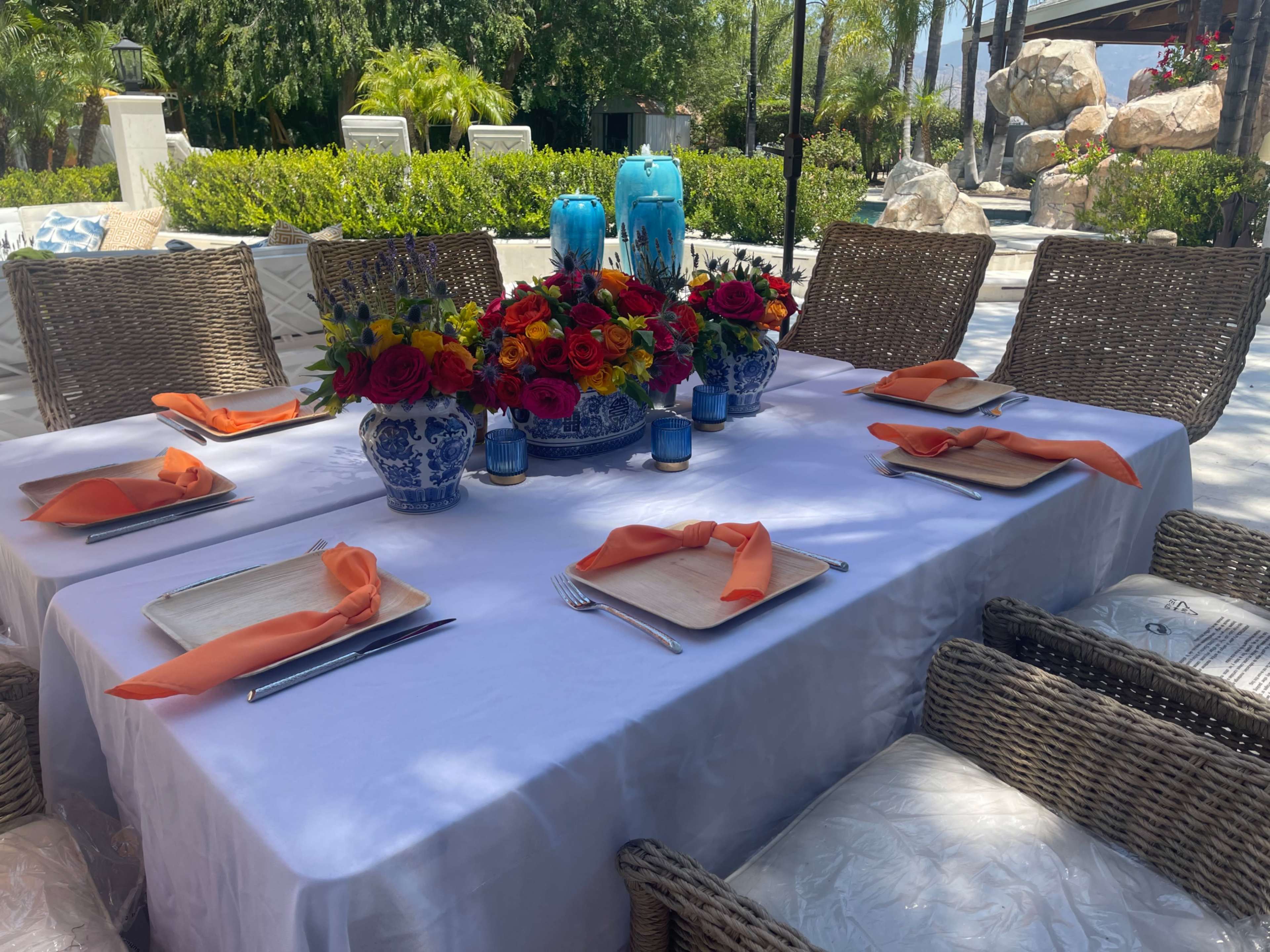 A dining table is set outdoors with an arrangement of colorful flowers, blue and white dishware, and orange napkins.