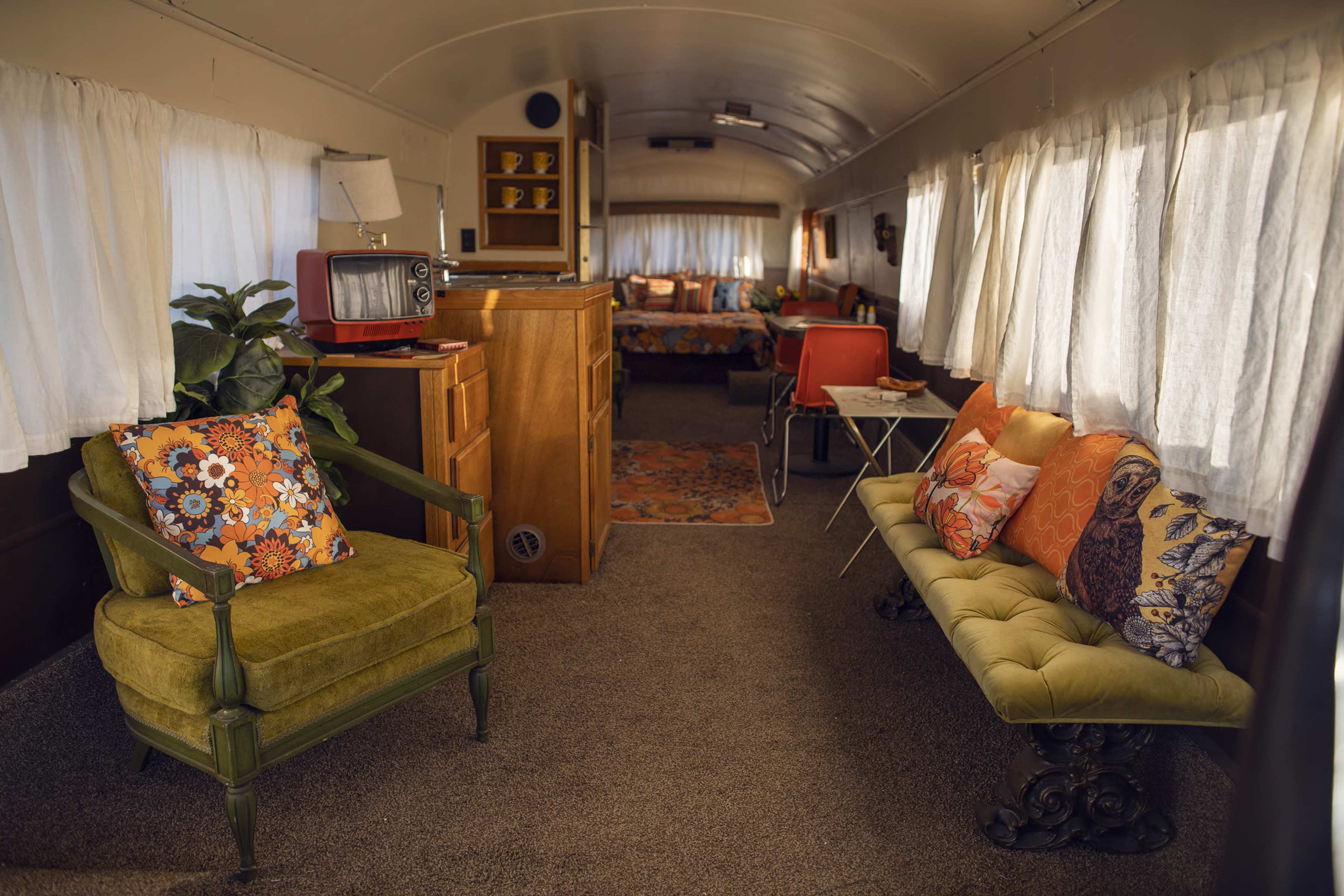 The interior of a vintage trailer features a cozy seating area with colorful throw pillows, a television, and a view into a bedroom space at the rear.