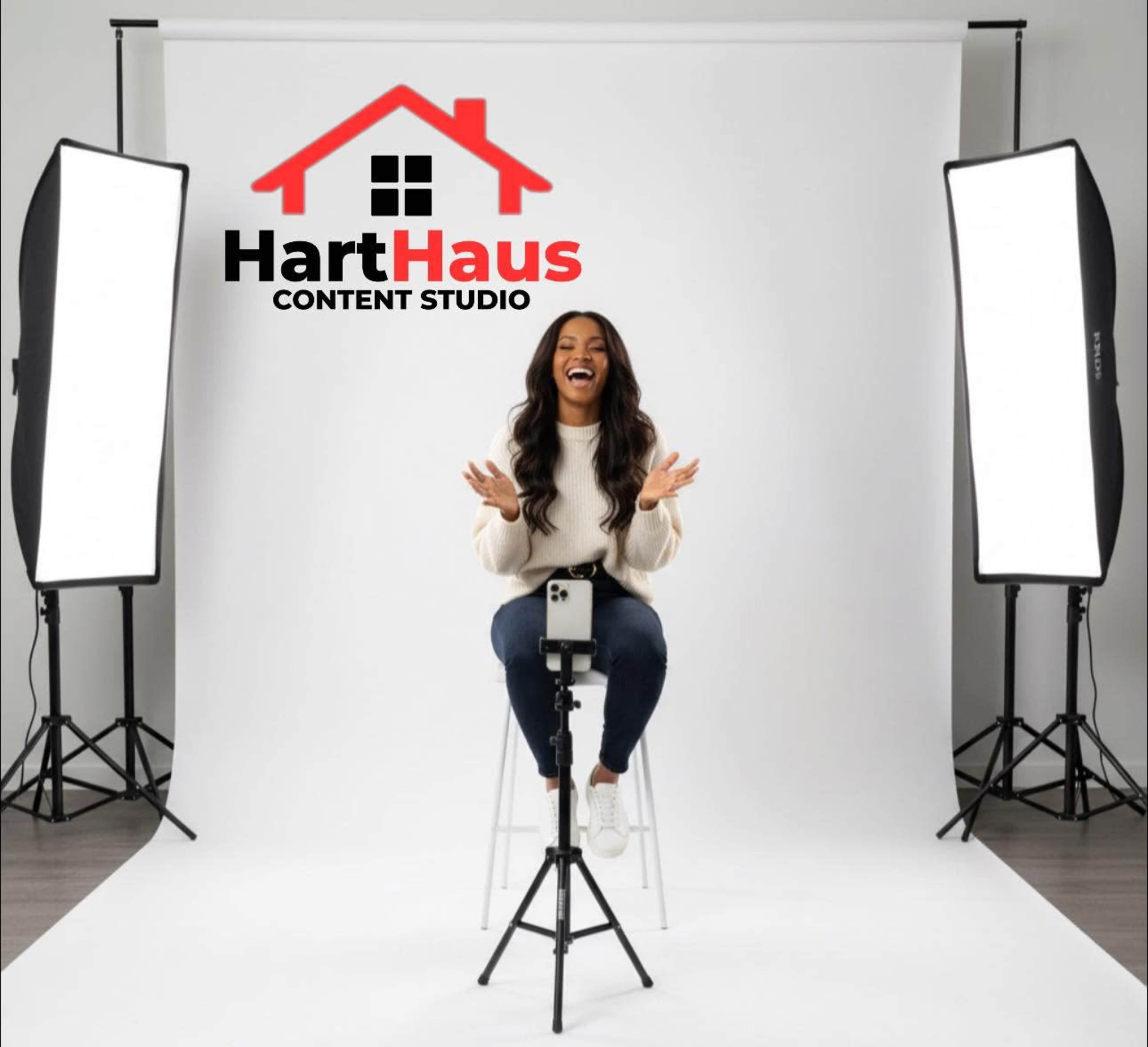 A woman sits on a stool in a brightly lit studio with white backdrops and lighting equipment, smiling and gesturing.