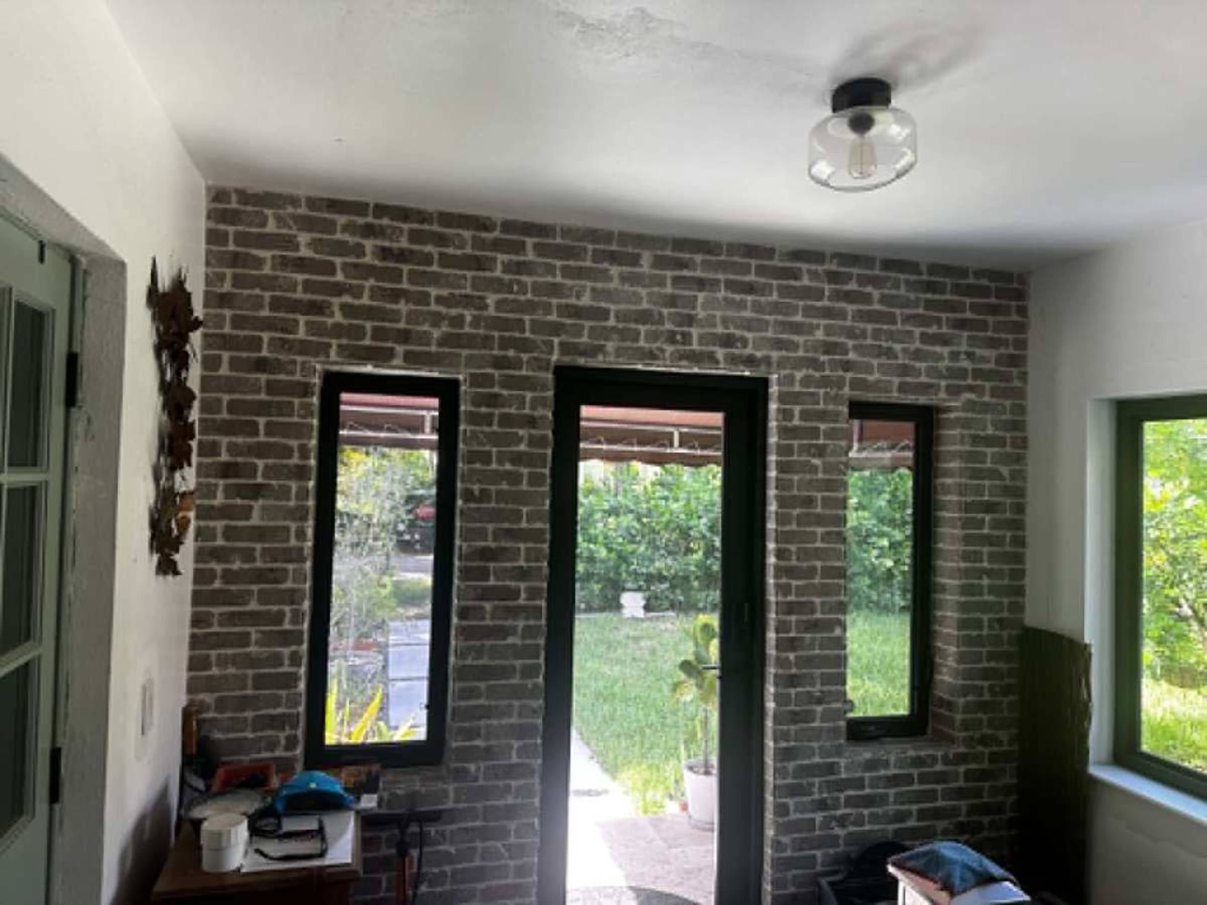 The image shows an interior space with brick-patterned walls, two large windows, and visible greenery outside.