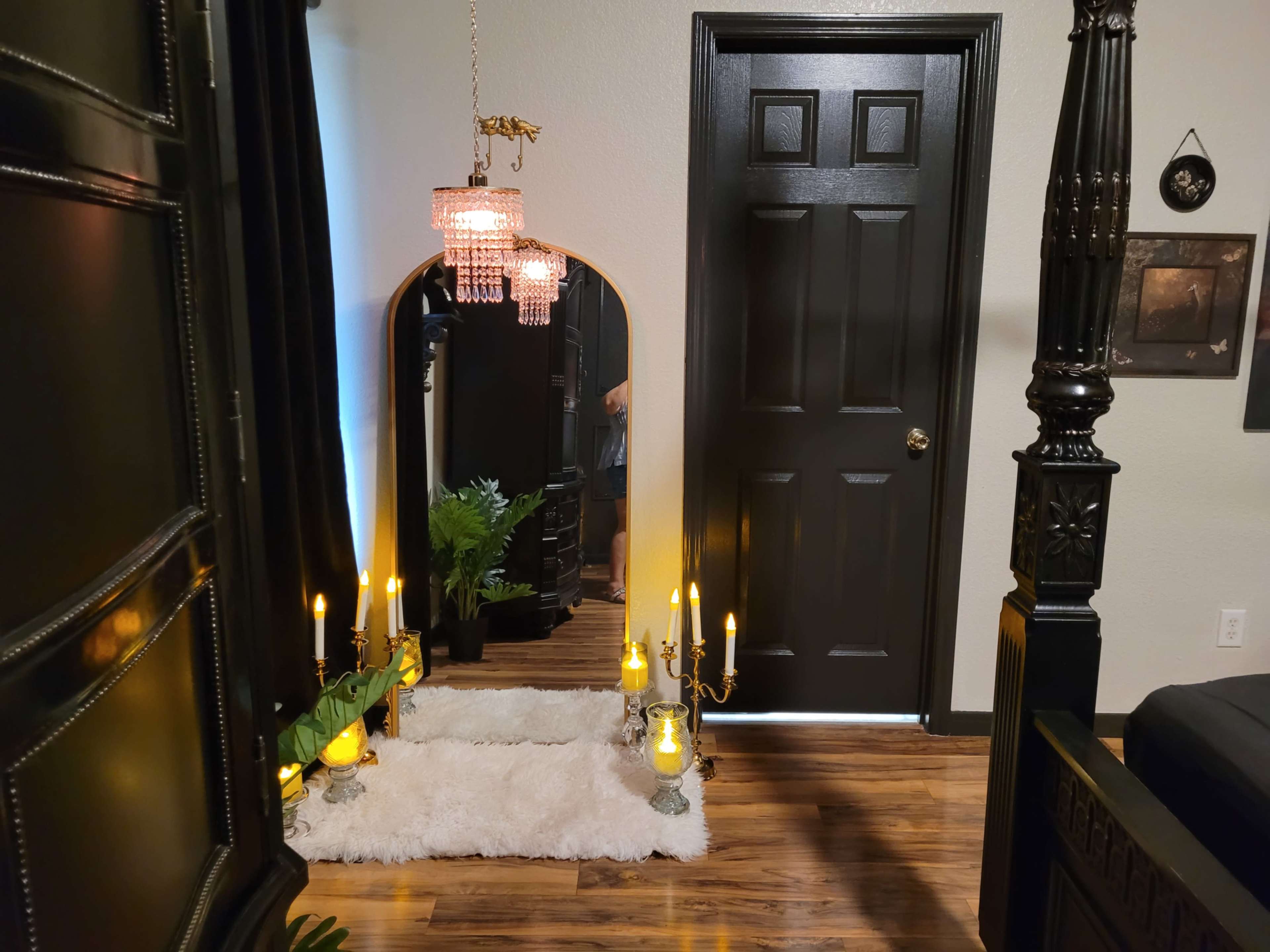 The scene features a stylish room with a large arched mirror, candles on a decorative stand, a plush white rug, and dark wooden flooring.