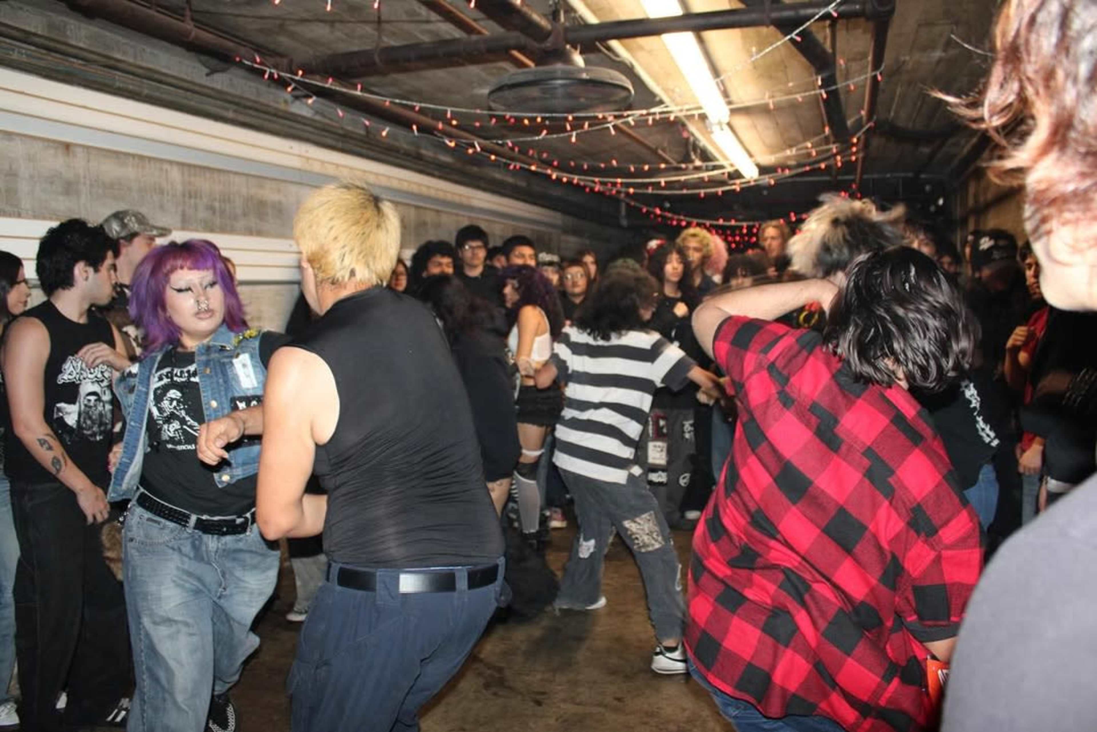 A crowd of young people is participating in a mosh pit in a dimly lit hallway decorated with string lights.
