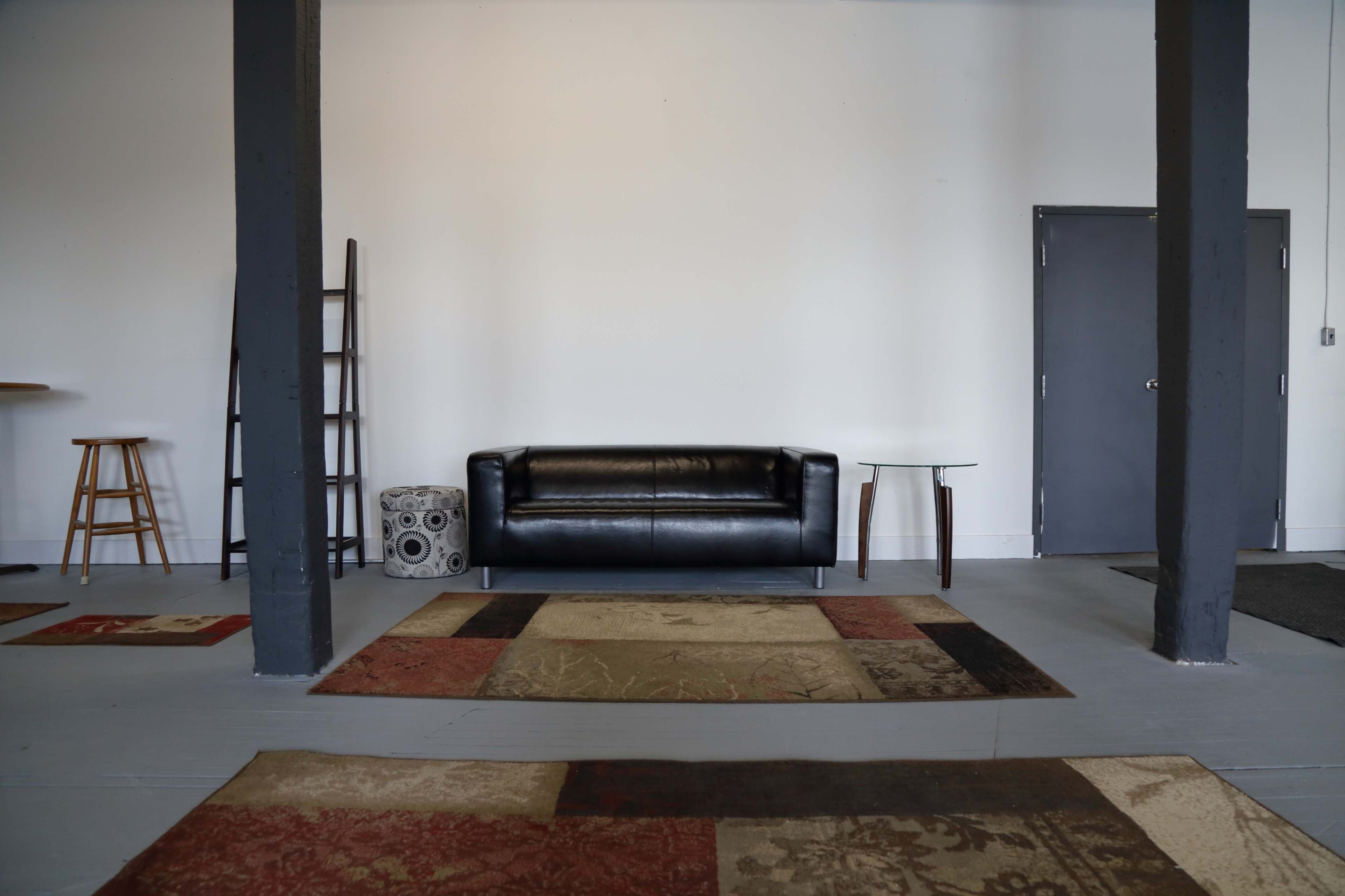 The scene shows a minimalist room with a black leather sofa, a wooden stool, a side table, and various area rugs on a gray floor.