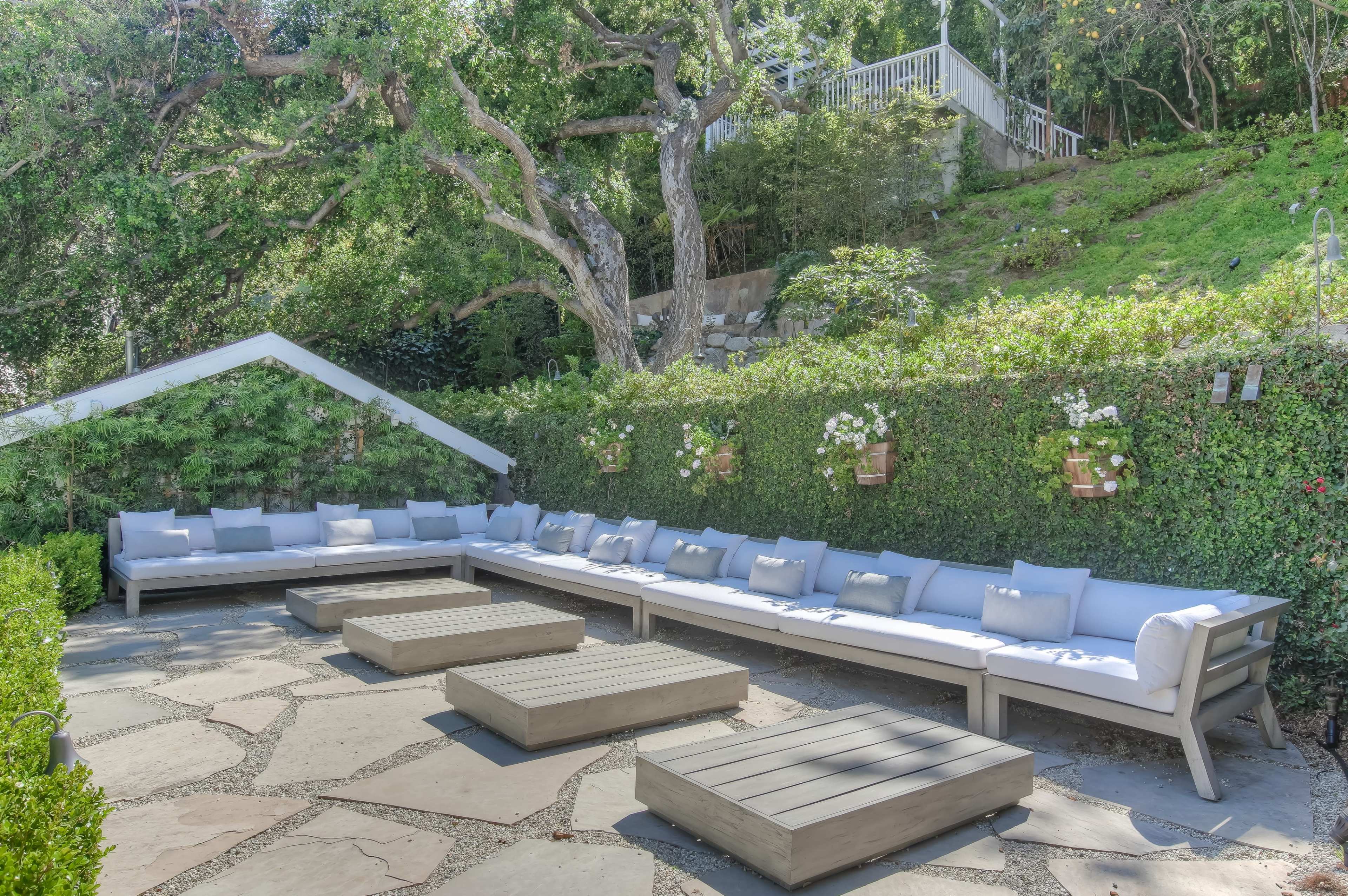 A spacious outdoor seating area with a long sectional couch and several low coffee tables arranged on a stone patio surrounded by greenery.