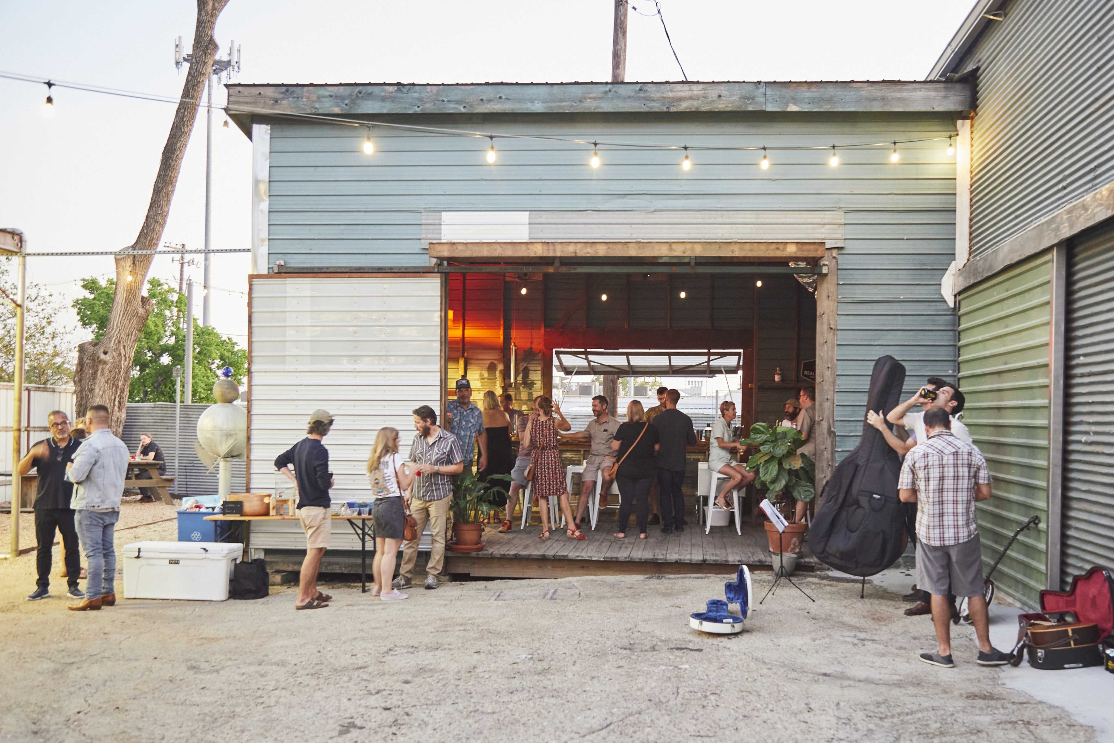 A gathering of people is taking place outside a rustic building with open doors, string lights overhead, and various seating arrangements.
