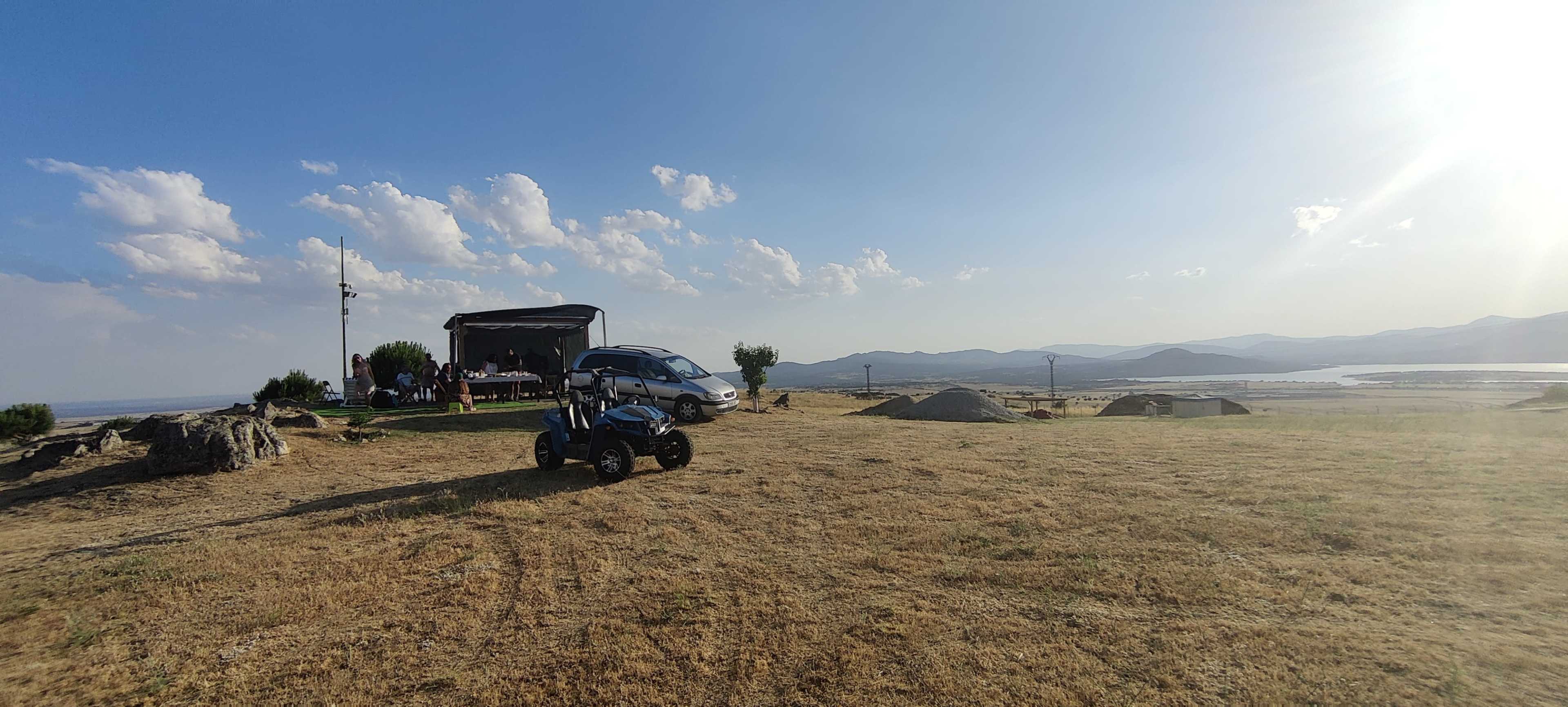 A gathering is taking place near a vehicle and an all-terrain vehicle on a grassy hillside overlooking a valley with distant mountains under a partly cloudy sky.