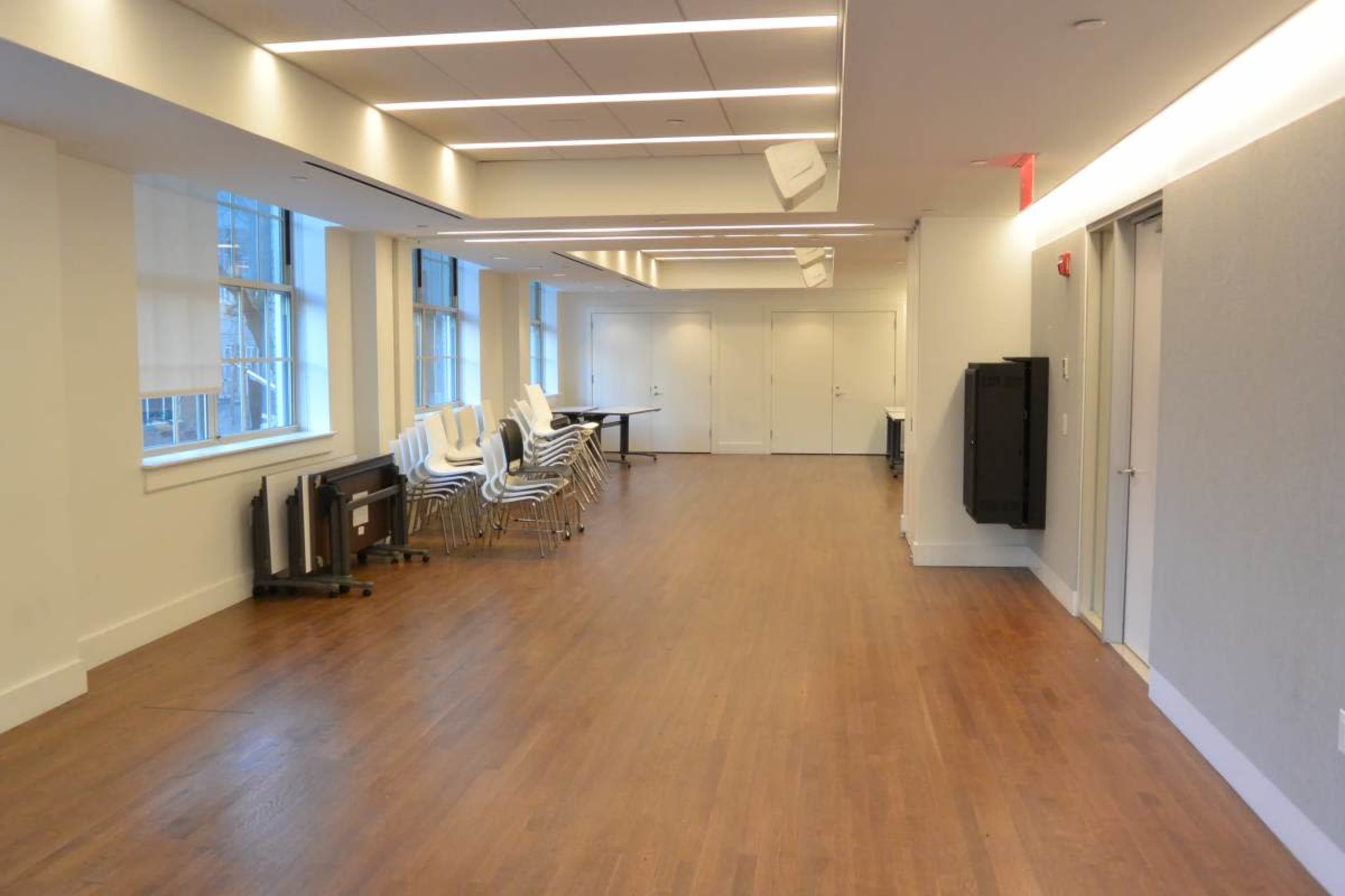 The image shows a brightly lit hallway with wooden flooring, featuring stacked chairs and a row of closed doors.