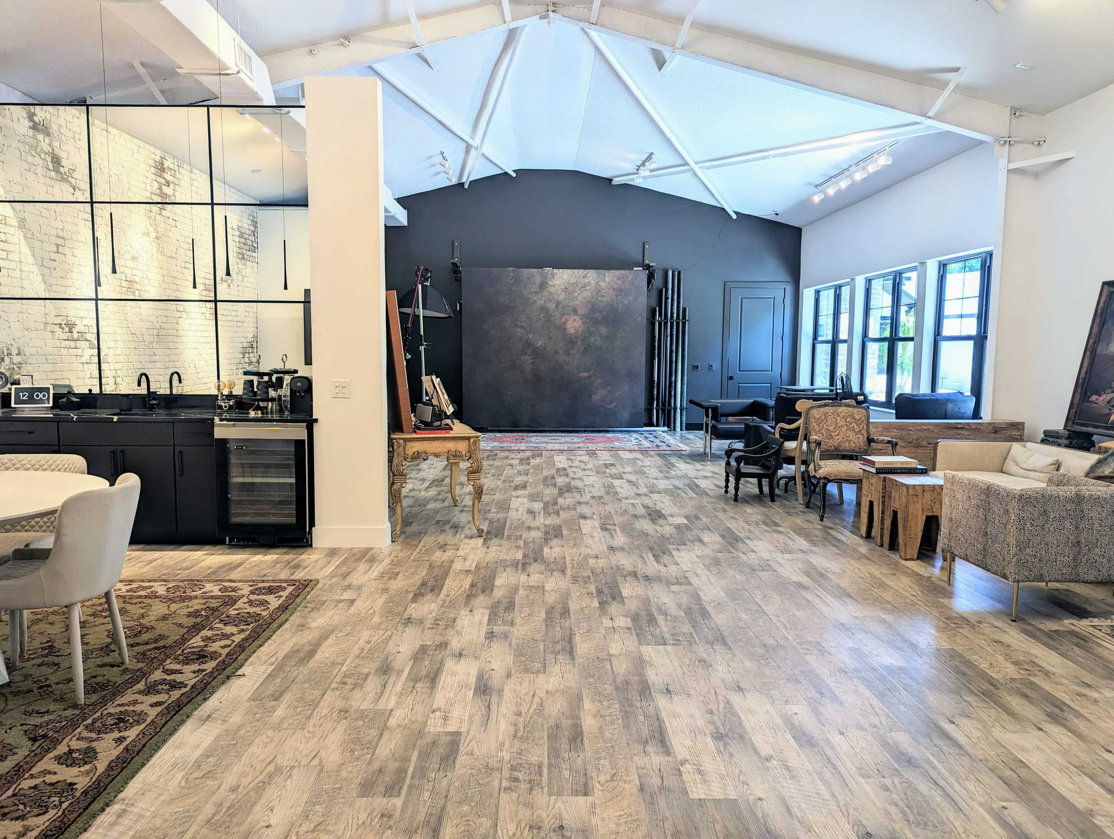 A spacious room with a modern kitchen area on one side and a stylish seating arrangement on the other, featuring a mix of furniture styles and large windows allowing natural light in.