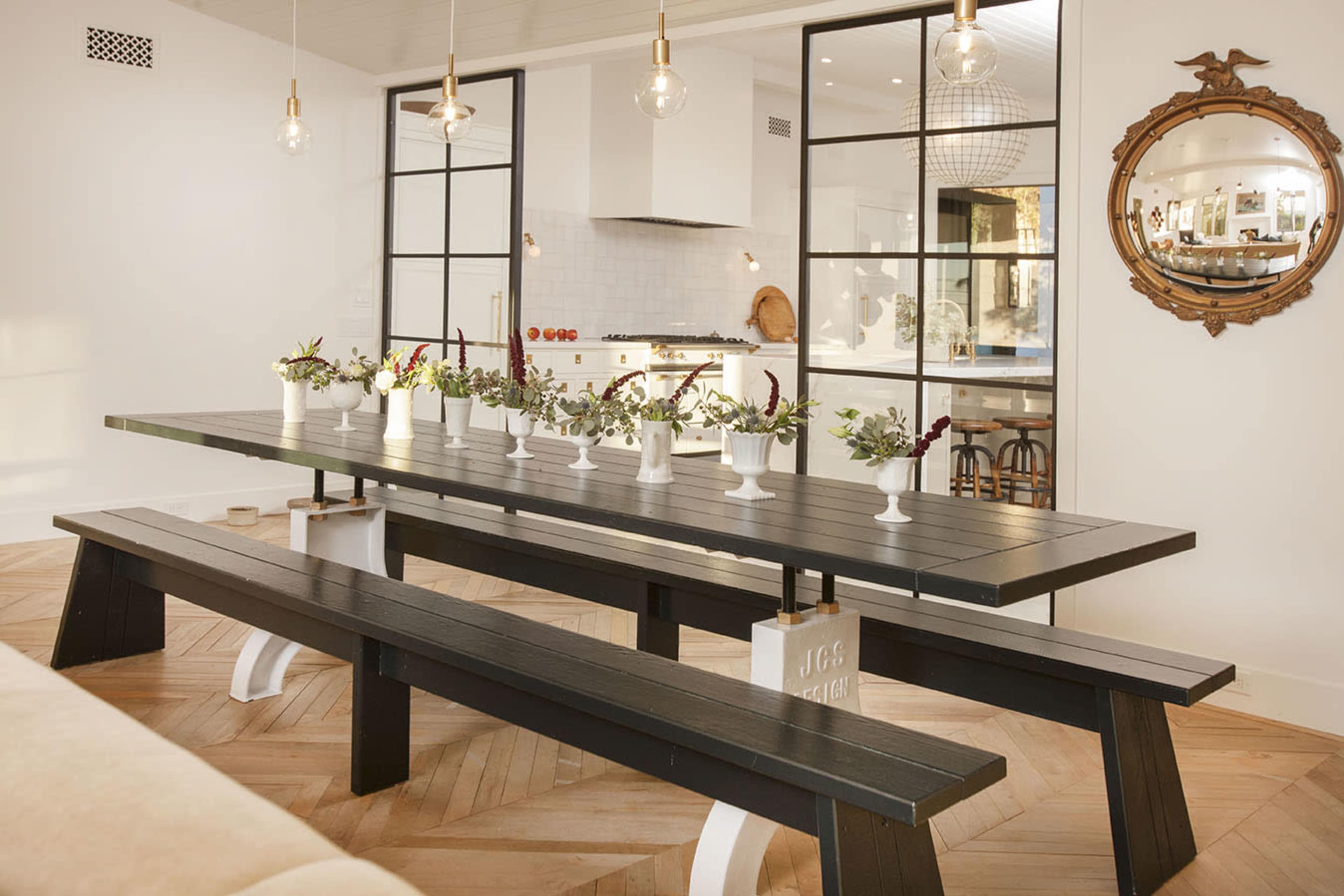 A long, dark wood dining table is set with small white vases of flowers, surrounded by matching benches in a bright, modern kitchen.