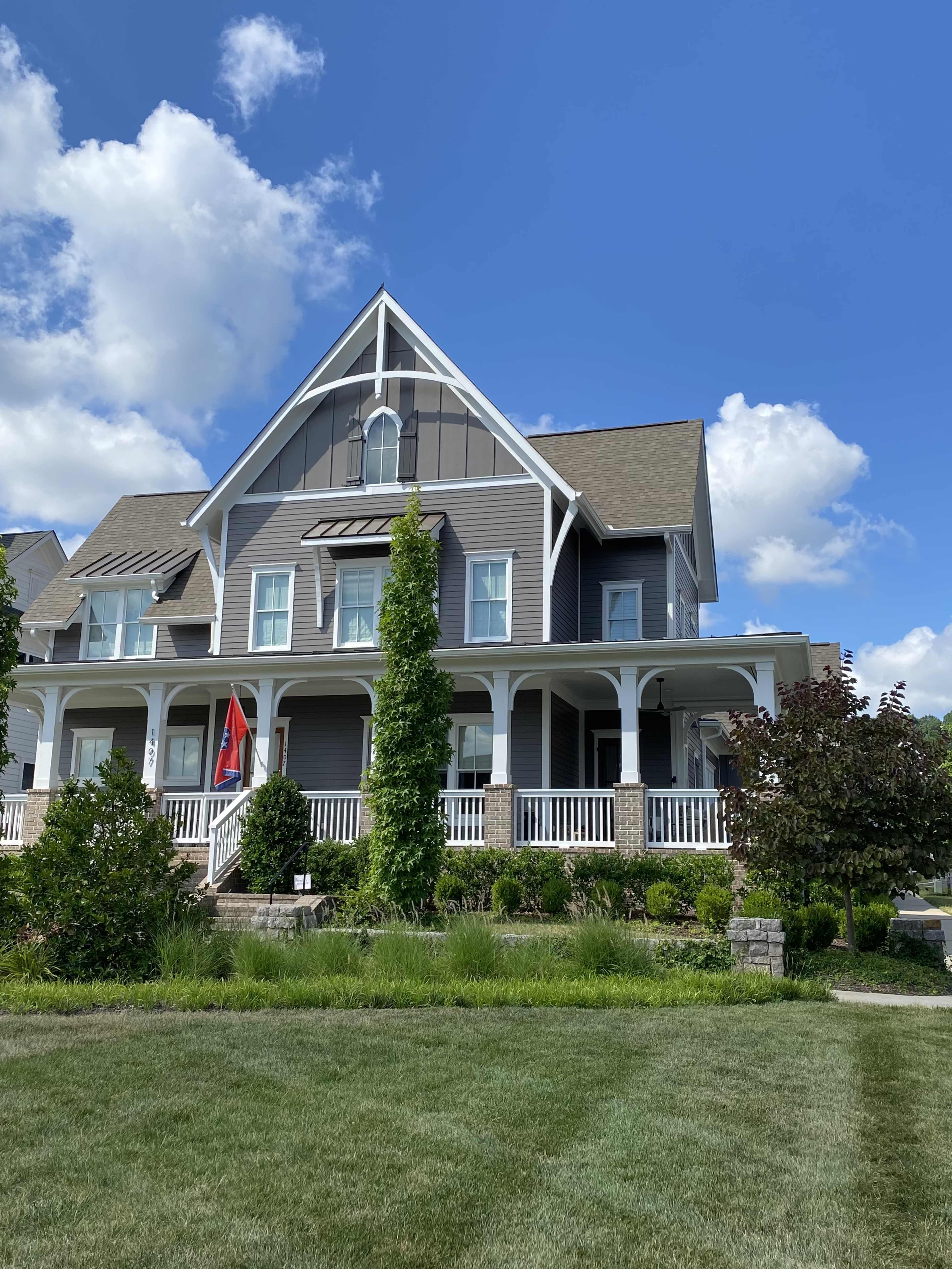 A large, two-story house with a gray exterior and white trim features an expansive porch and landscaped front yard under a blue sky with clouds.