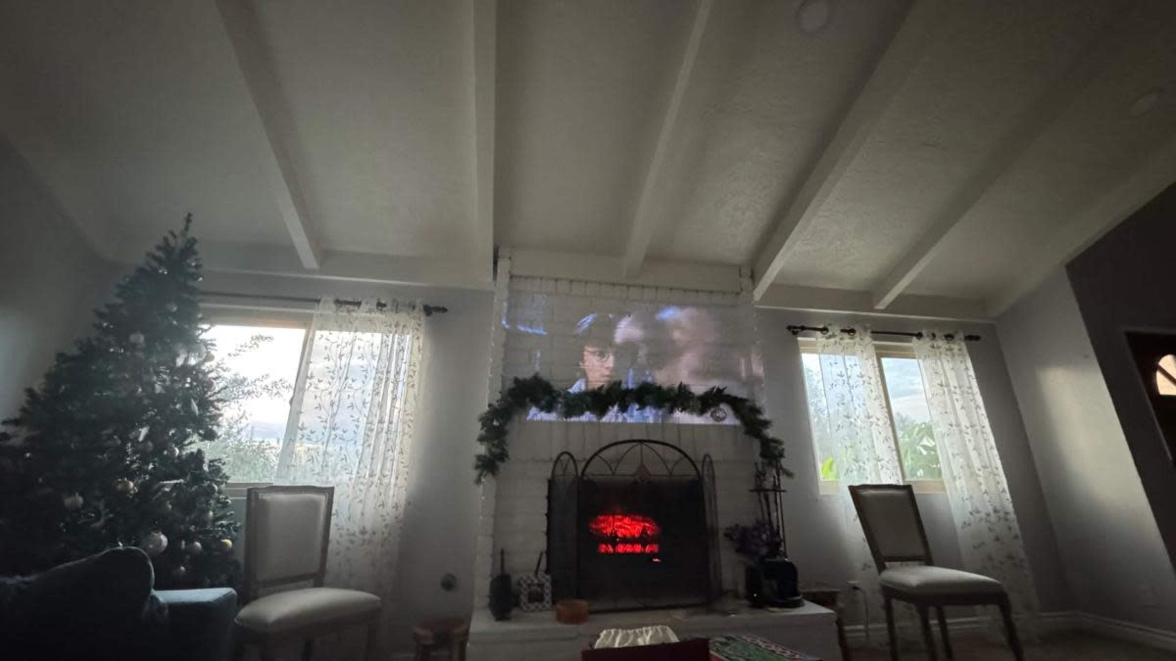 A projector displays a movie on a white wall in a living room decorated with a Christmas tree and a fireplace.