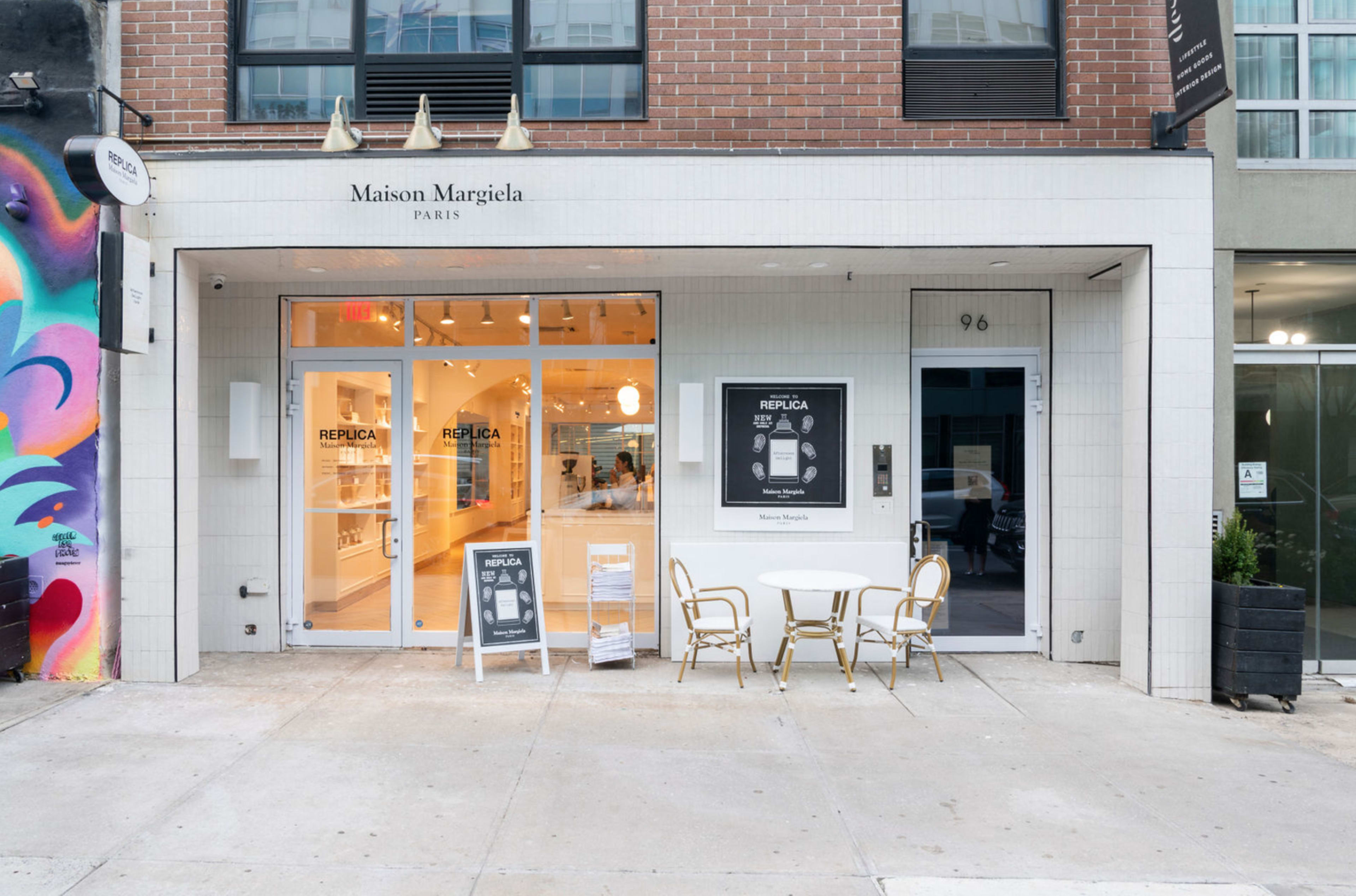 The image shows the exterior of a Maison Margiela boutique with large glass windows, a white tiled facade, and outdoor seating featuring a small round table and chairs.