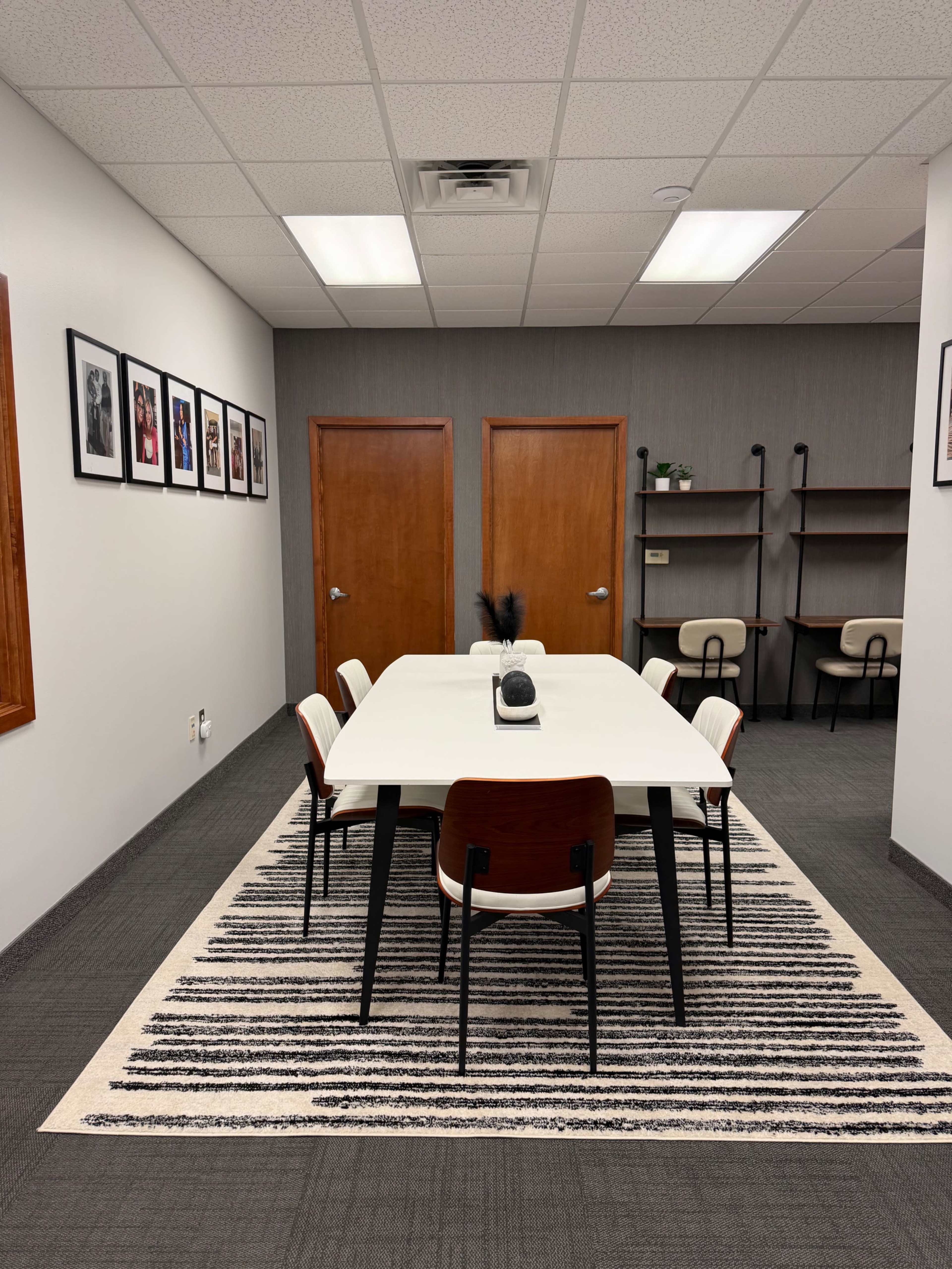 The image shows a modern conference room with a rectangular white table surrounded by four chairs, a striped area rug, and framed photos on the walls.