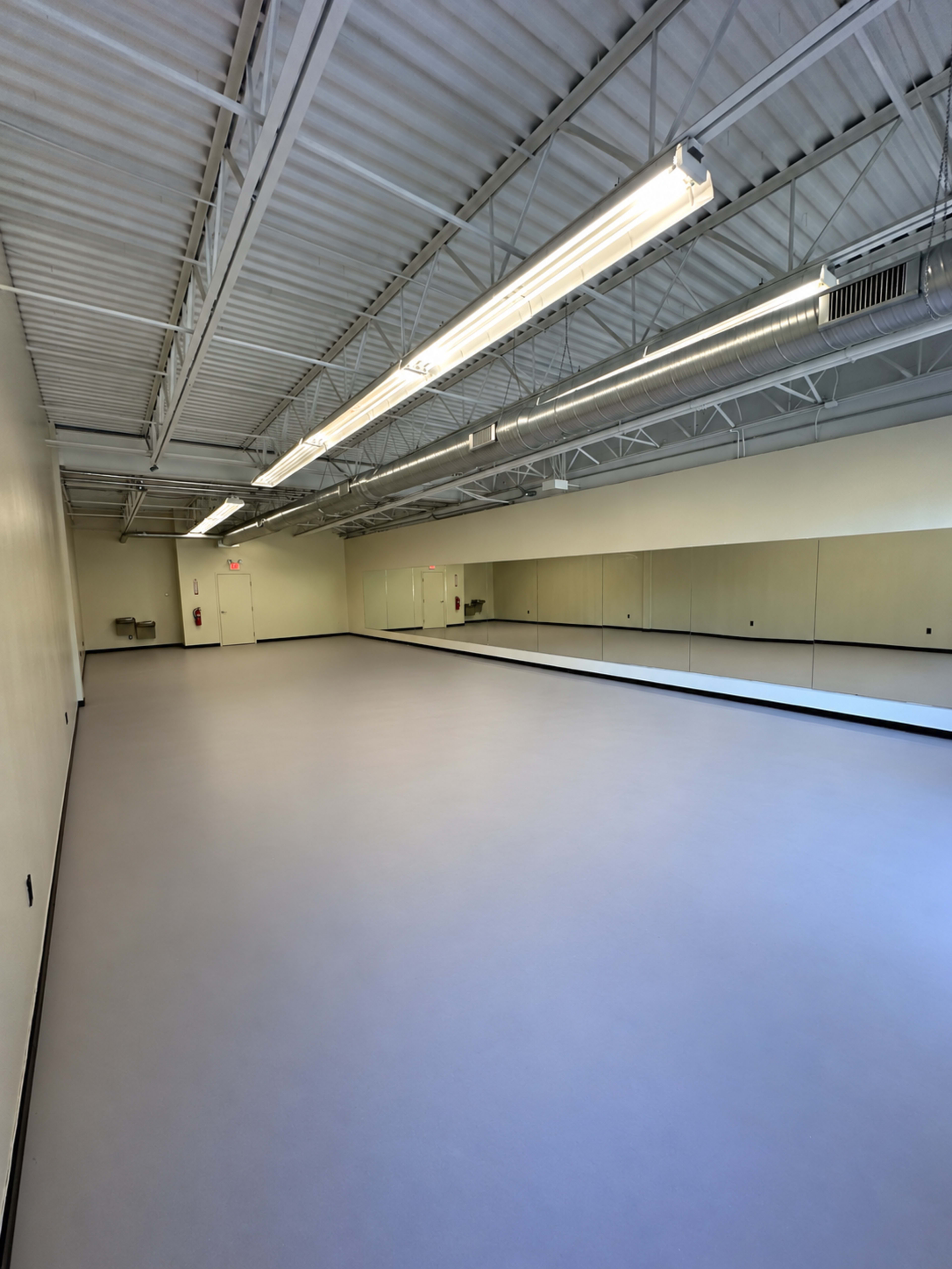 The image shows a spacious, empty dance studio with large mirrors along one wall and bright overhead lighting.