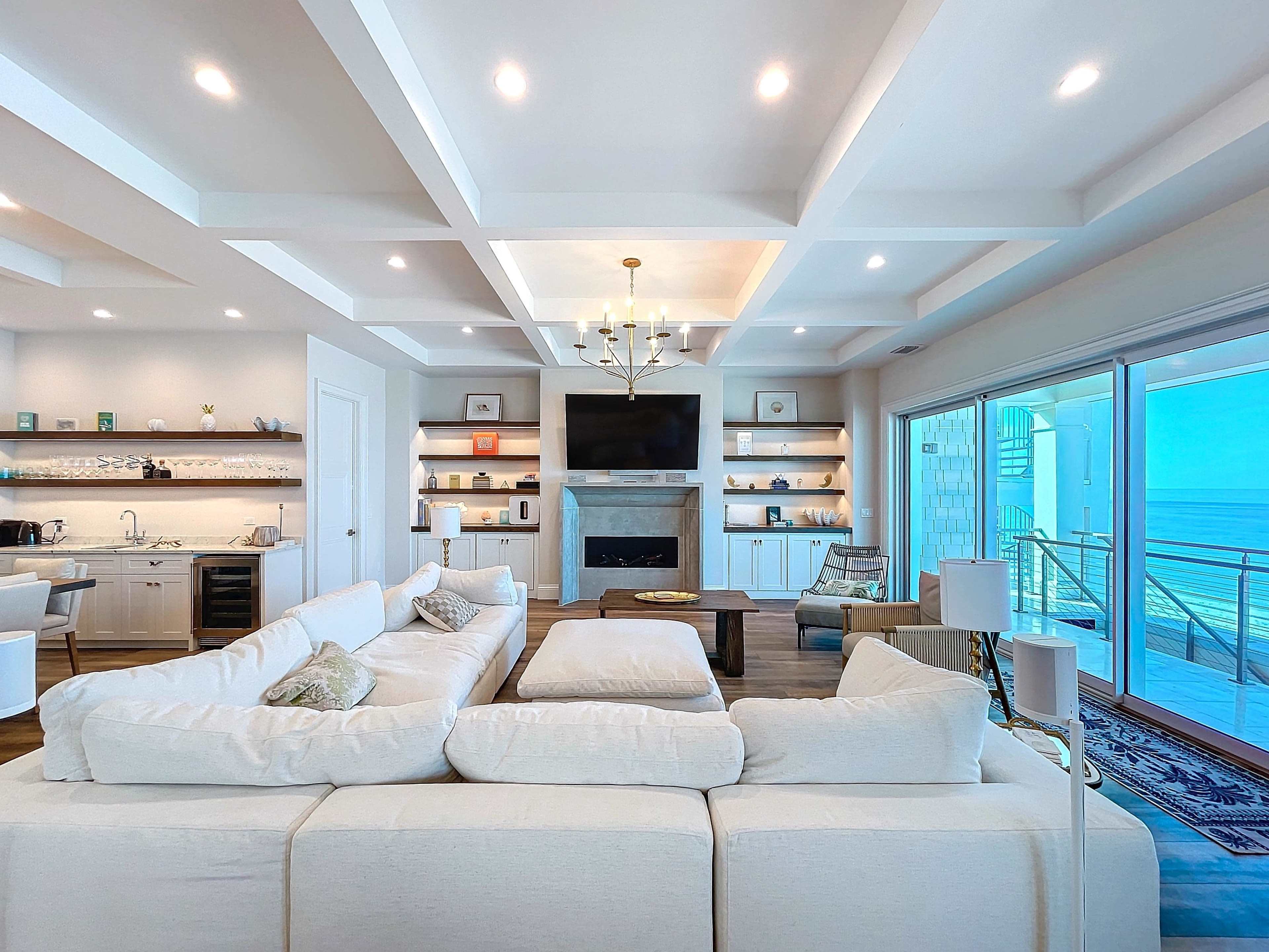 The image shows a modern living room with a large white sectional sofa, a coffee table, and a view of the ocean through sliding glass doors.