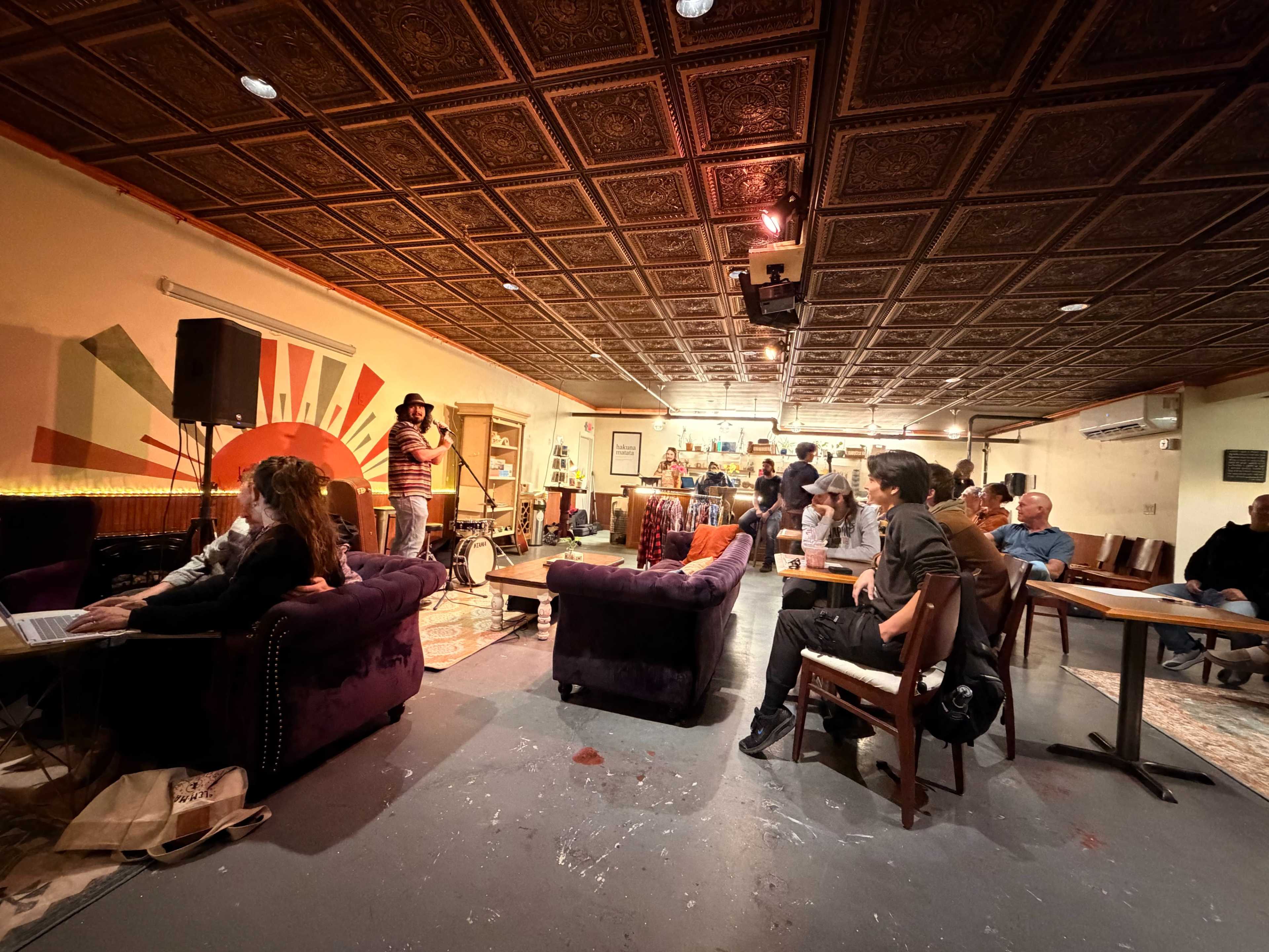 The image shows a lively café or lounge with guests seated at tables and couches while a performer stands on a small stage in the background.