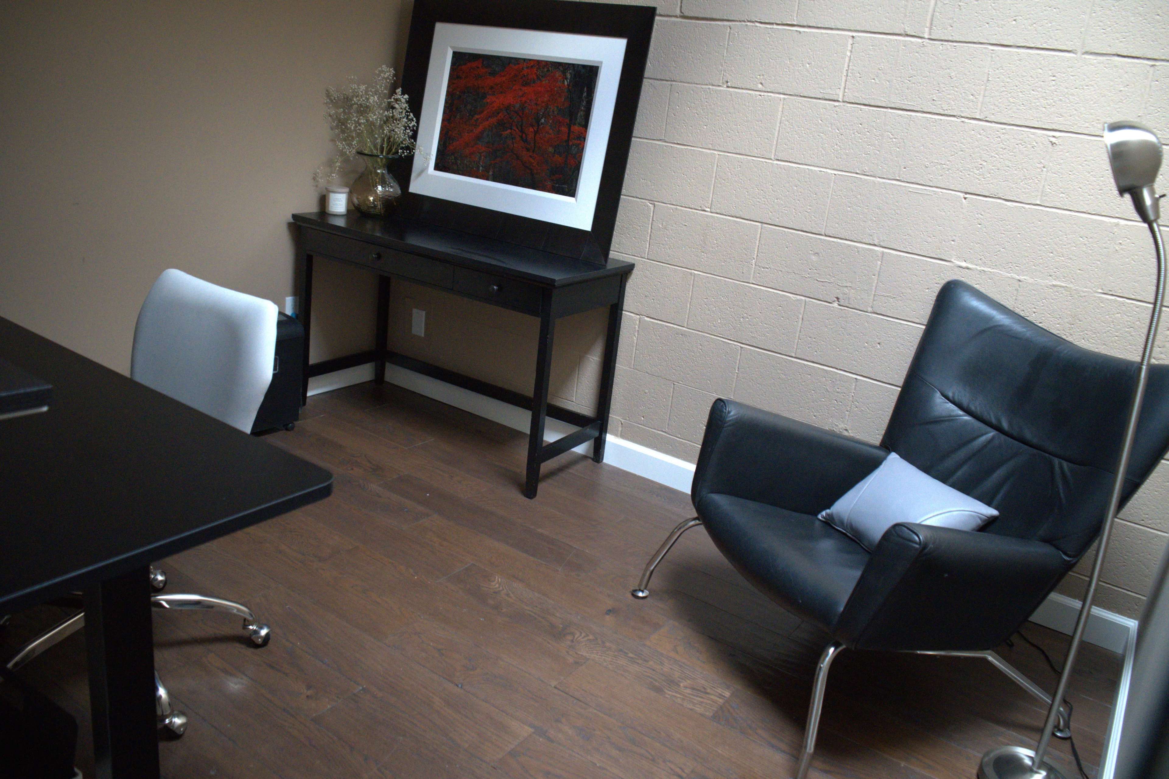 The image shows a minimalist office space with a black chair, a desk, and a framed piece of art on the wall.