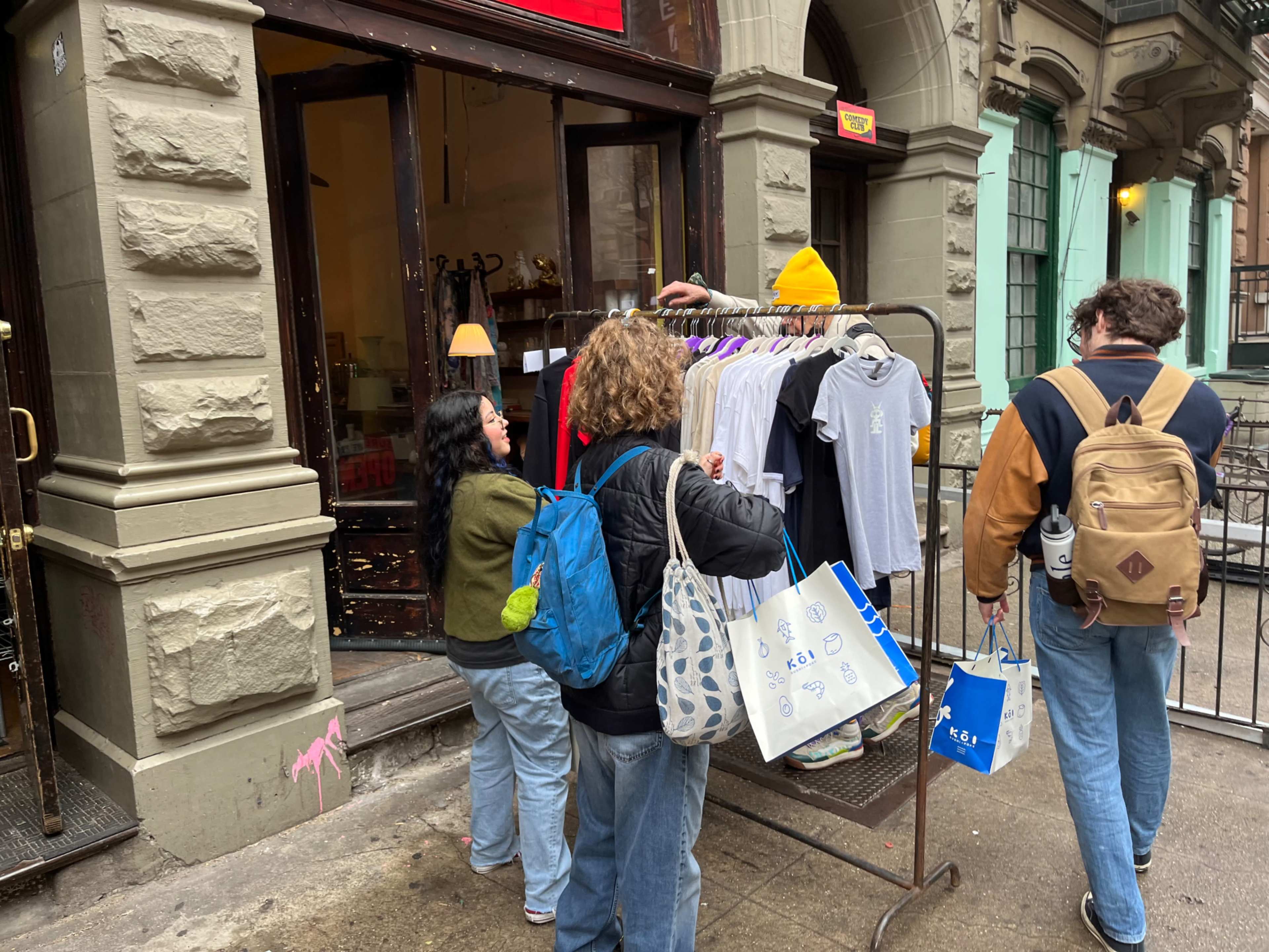 A small group of people browses a rack of clothing outside a shop in a city setting.