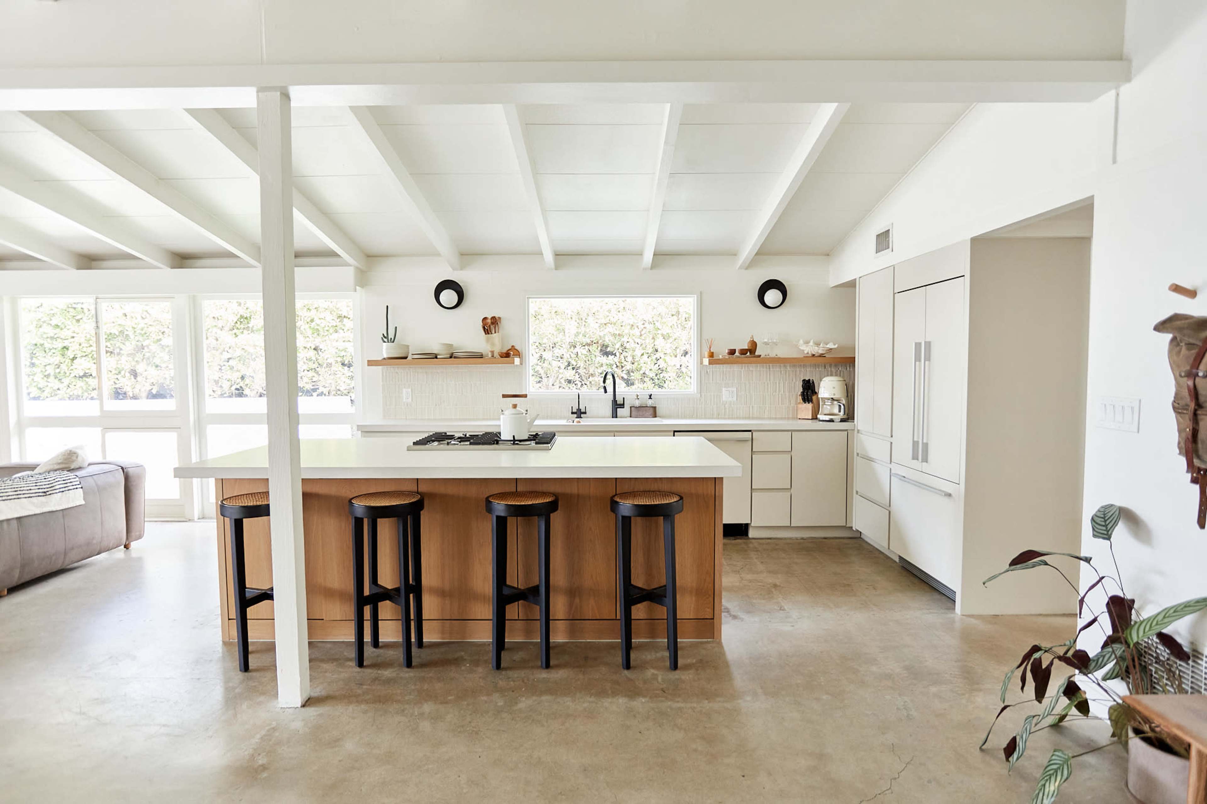 A modern kitchen features a central island with bar stools, white cabinetry, and large windows allowing natural light.
