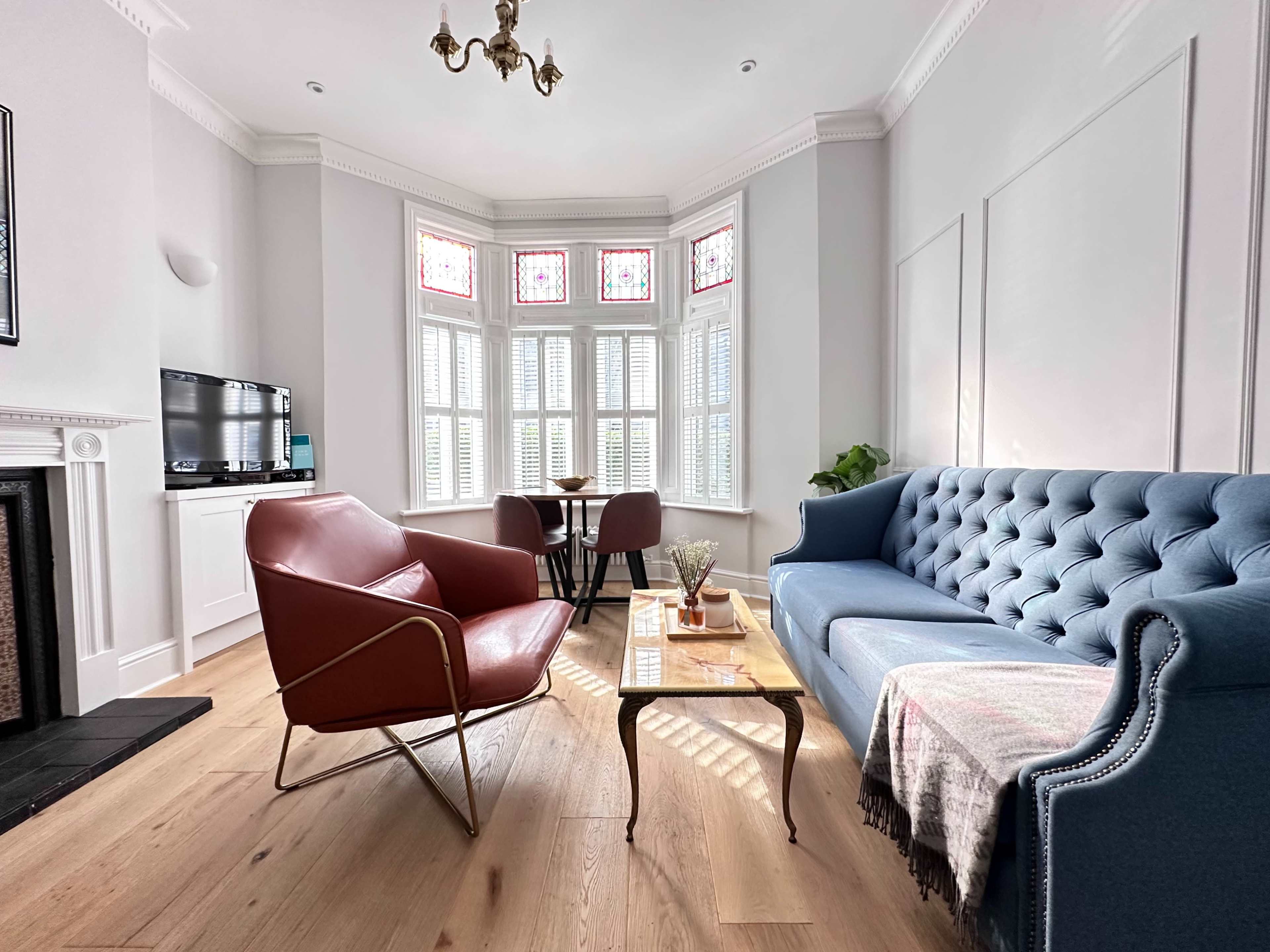 A brightly lit living room featuring a blue tufted sofa, a red armchair, a wooden coffee table, and a small dining area with a round table and chairs, all set against a backdrop of large windows.