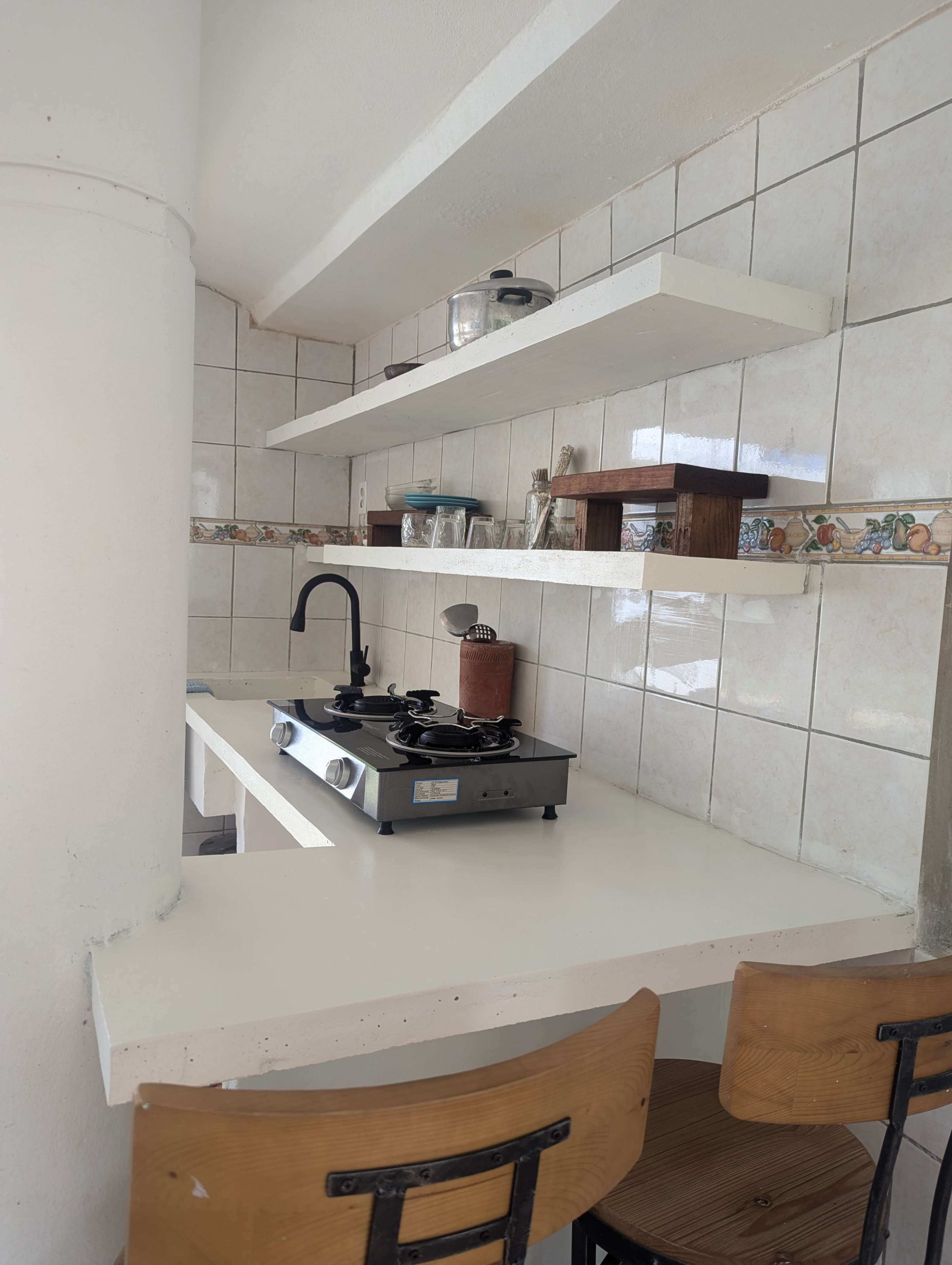 The image shows a small kitchen area with a gas stove, two shelves above, and a couple of wooden stools at a counter.