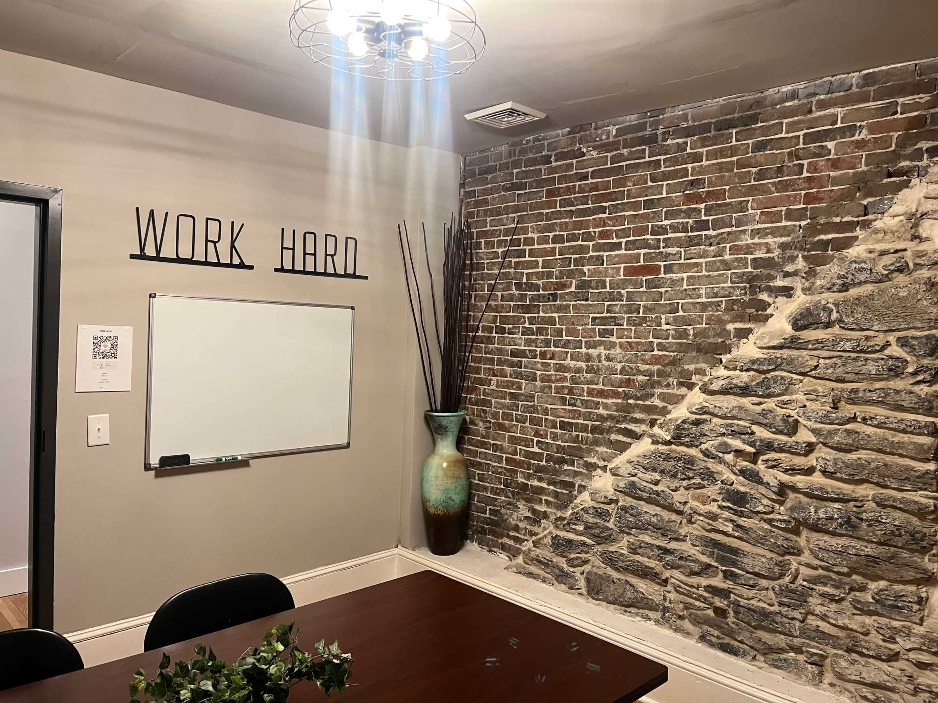 The image shows a meeting room with a wooden table, a whiteboard on the wall, a decorative vase, and an exposed brick feature.