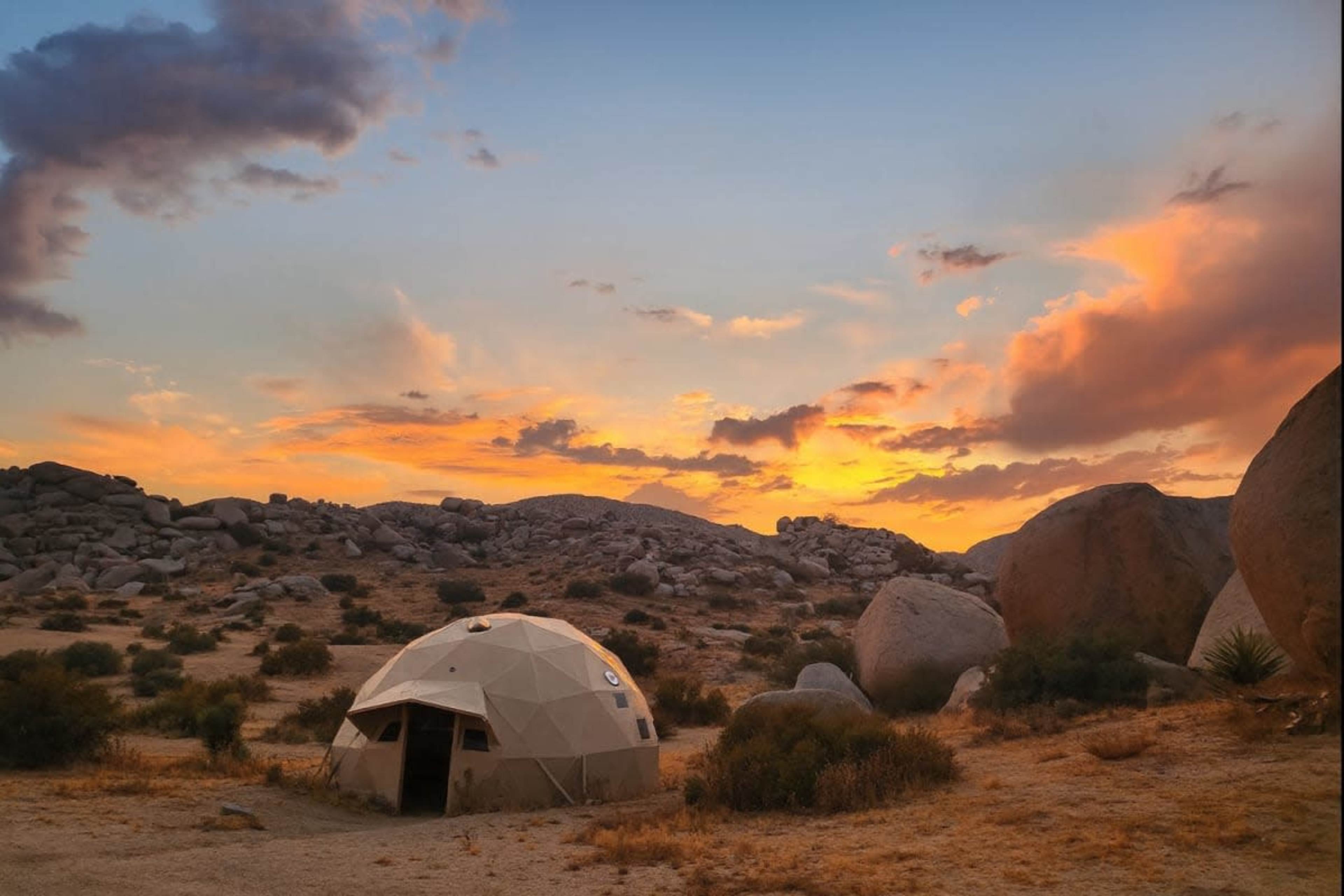A geodesic dome is set against a desert landscape with large boulders and a vibrant sunset sky.