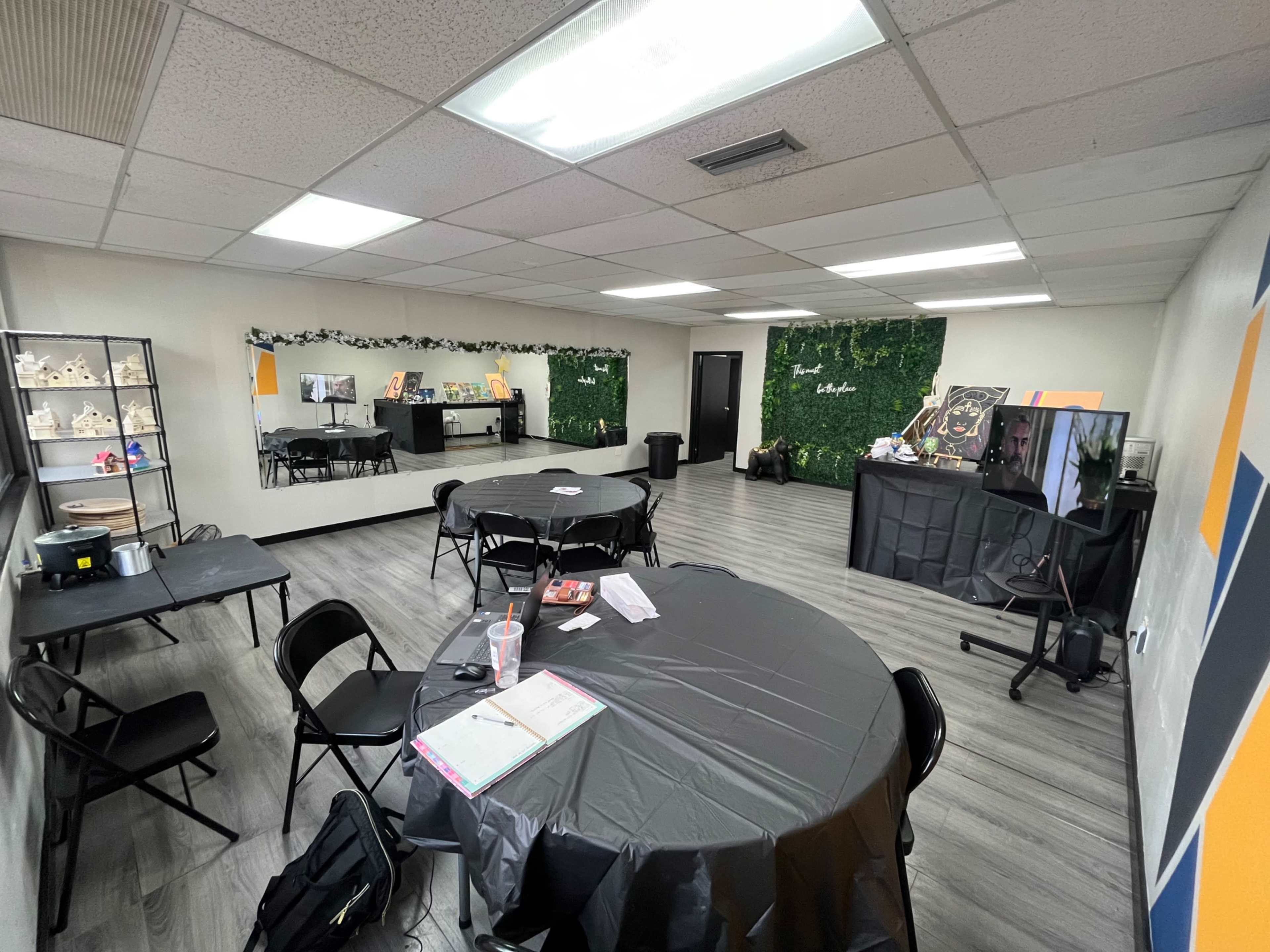 An empty meeting room with several black round tables, a mirrored wall, and a green plant backdrop.