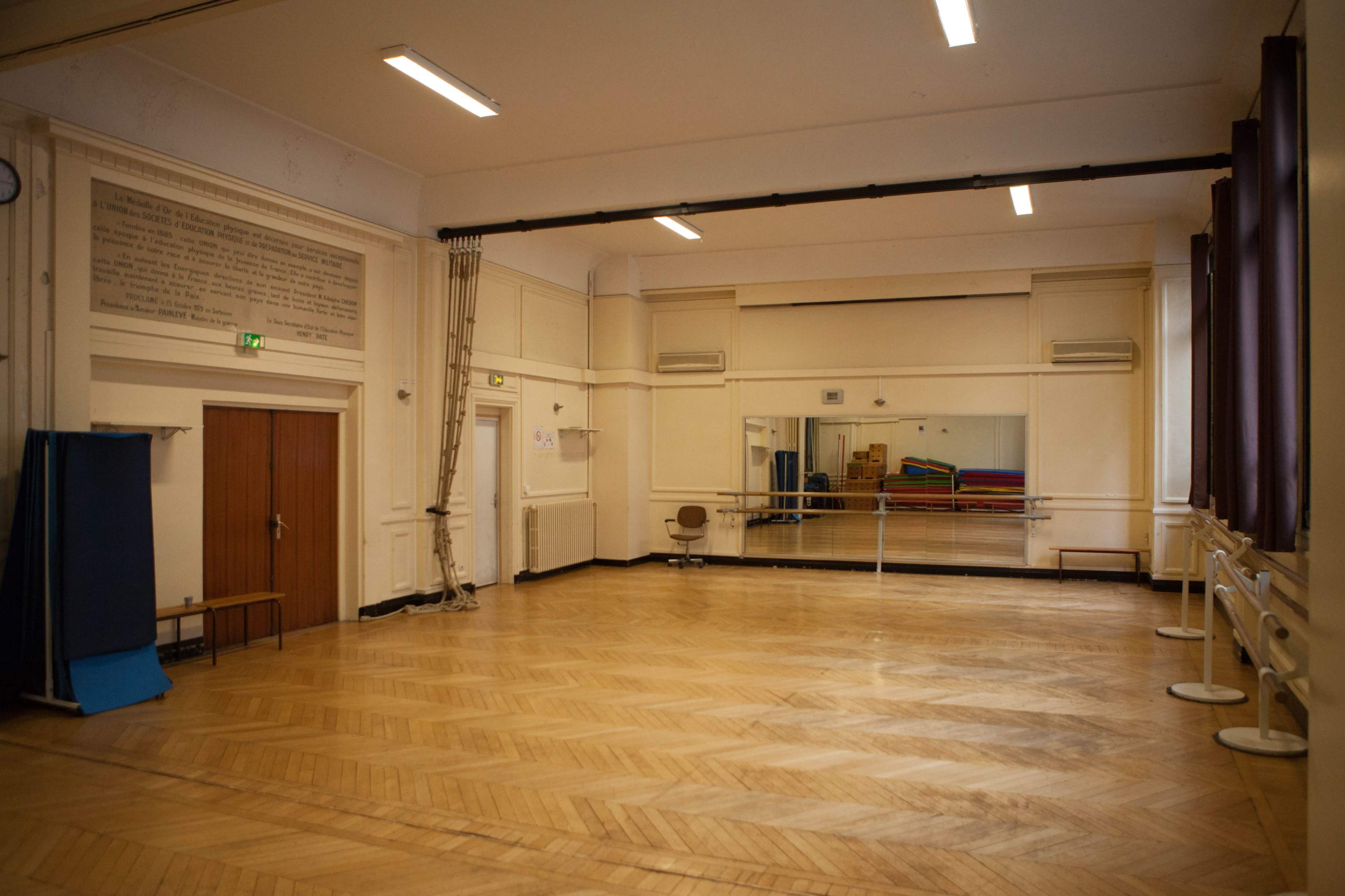 The image shows a spacious, empty room with wooden flooring, large mirrors on one wall, and exercise mats stacked in the corner.
