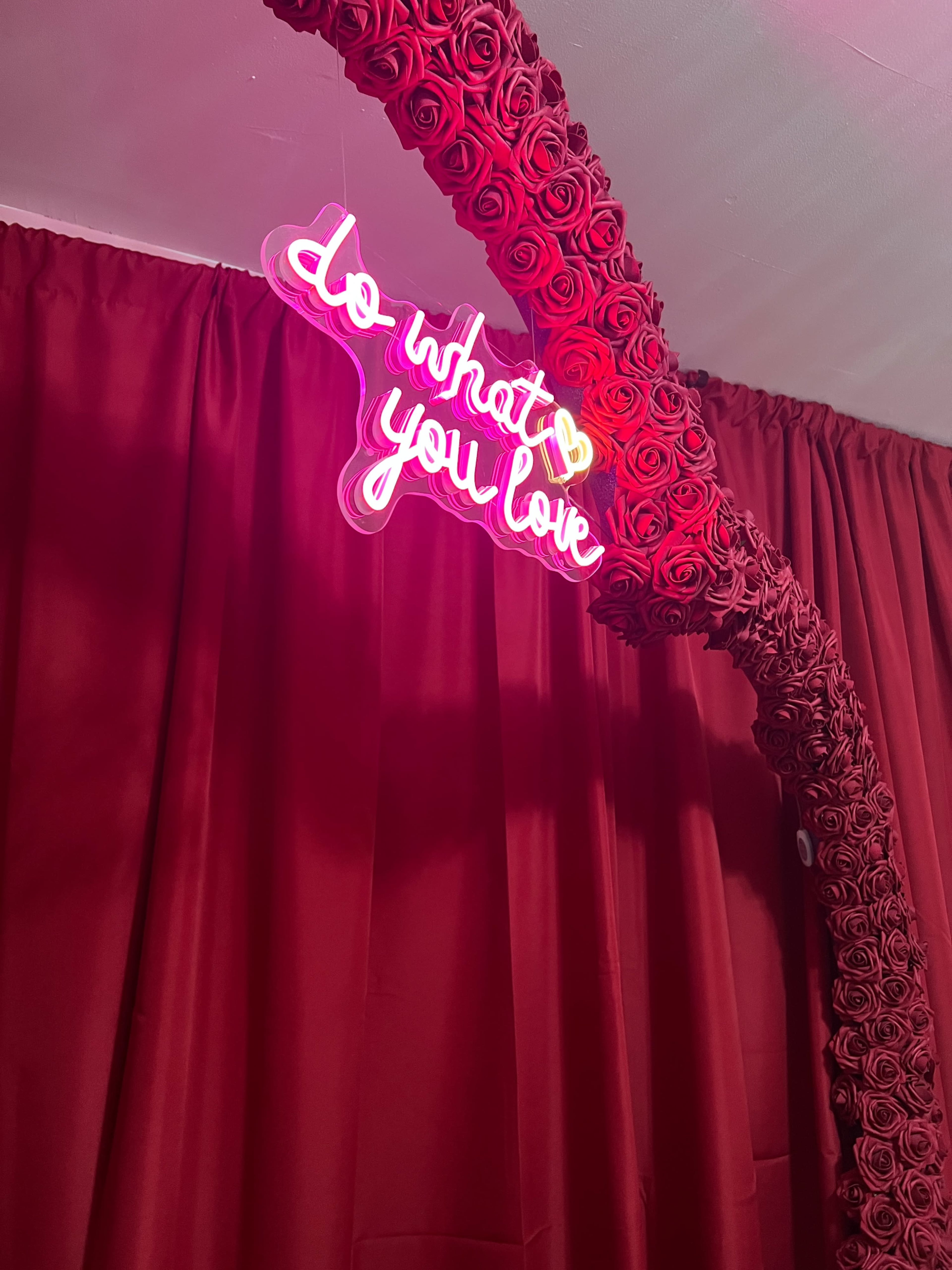A neon sign saying "do what you love" is suspended from an arch covered in red roses, in front of a backdrop of red curtains.