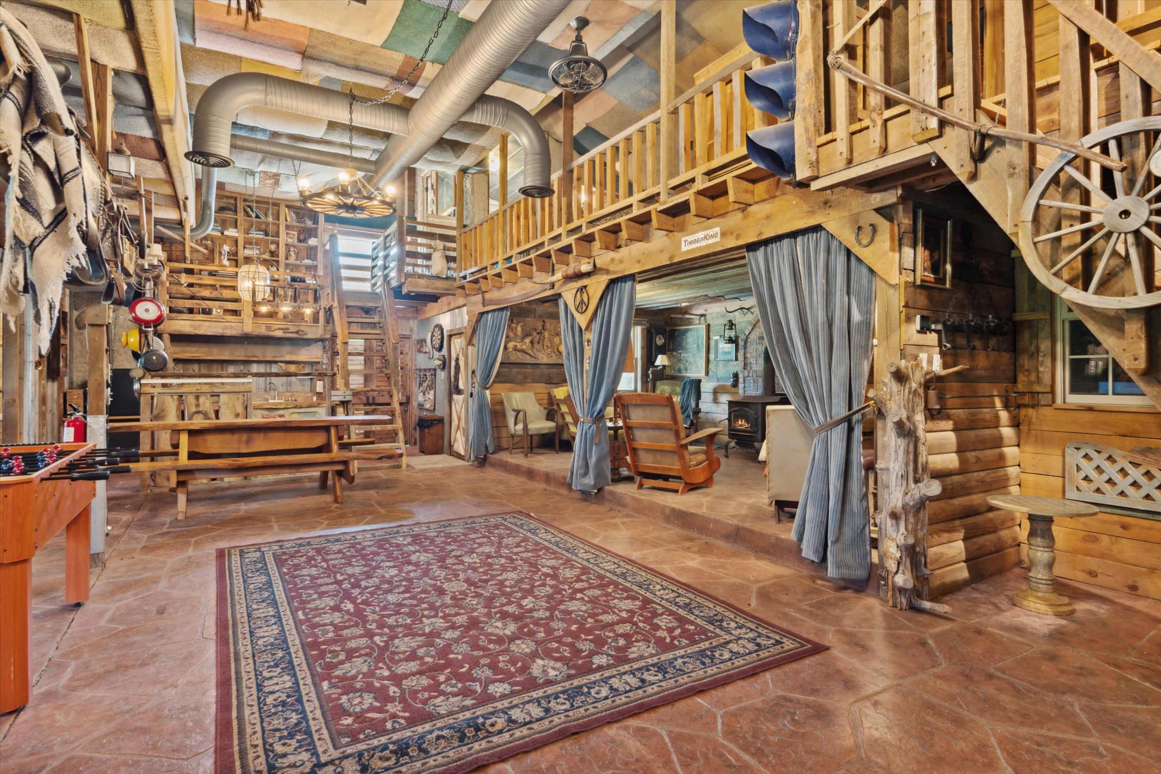 The image shows a spacious, rustic interior with wooden beams, a staircase leading to an upper level, and various furniture pieces arranged around a decorative rug.